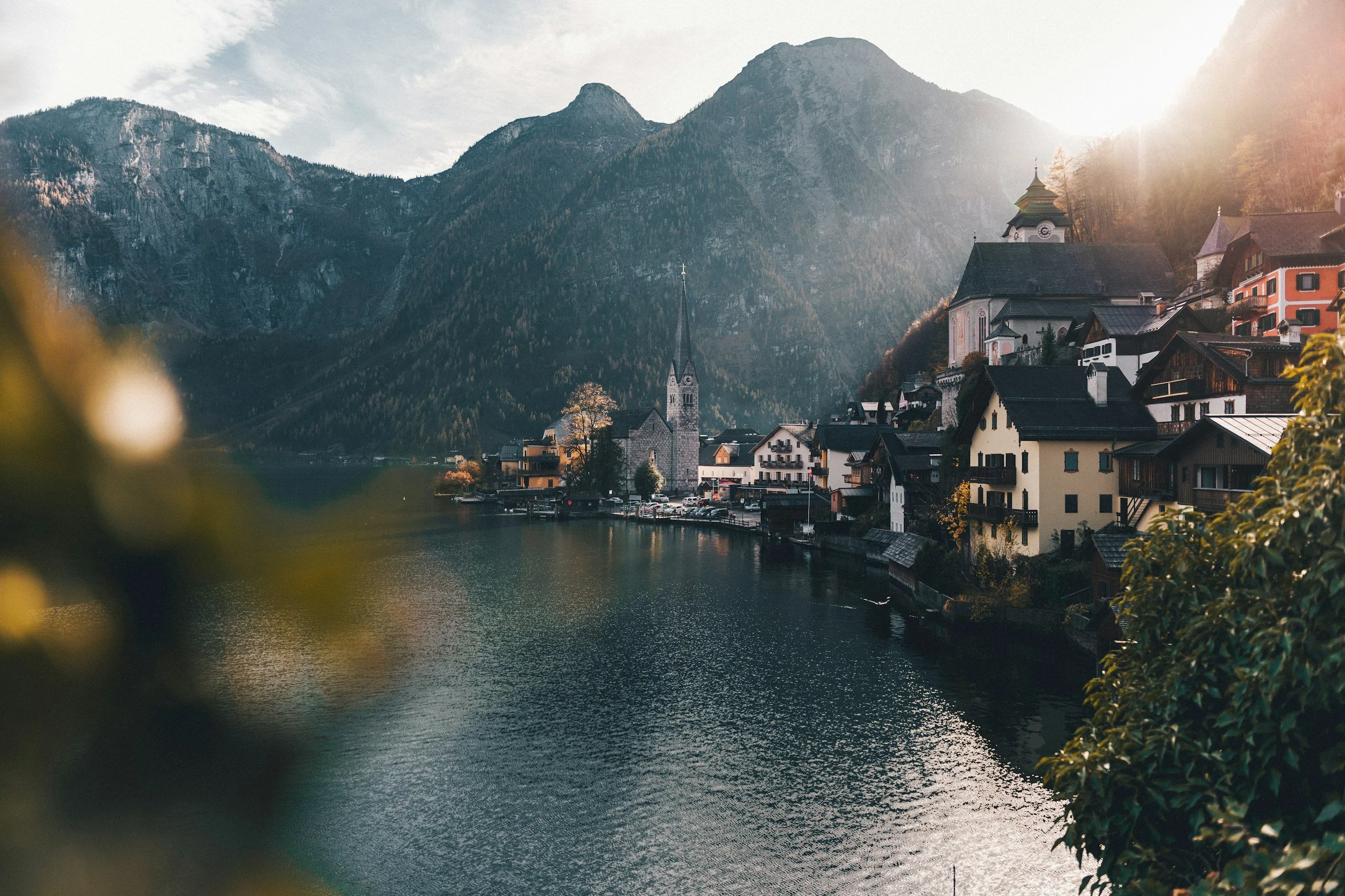 A scenic lakeside village with colorful houses and church steeples, surrounded by tall mountains and partially illuminated by sunlight.