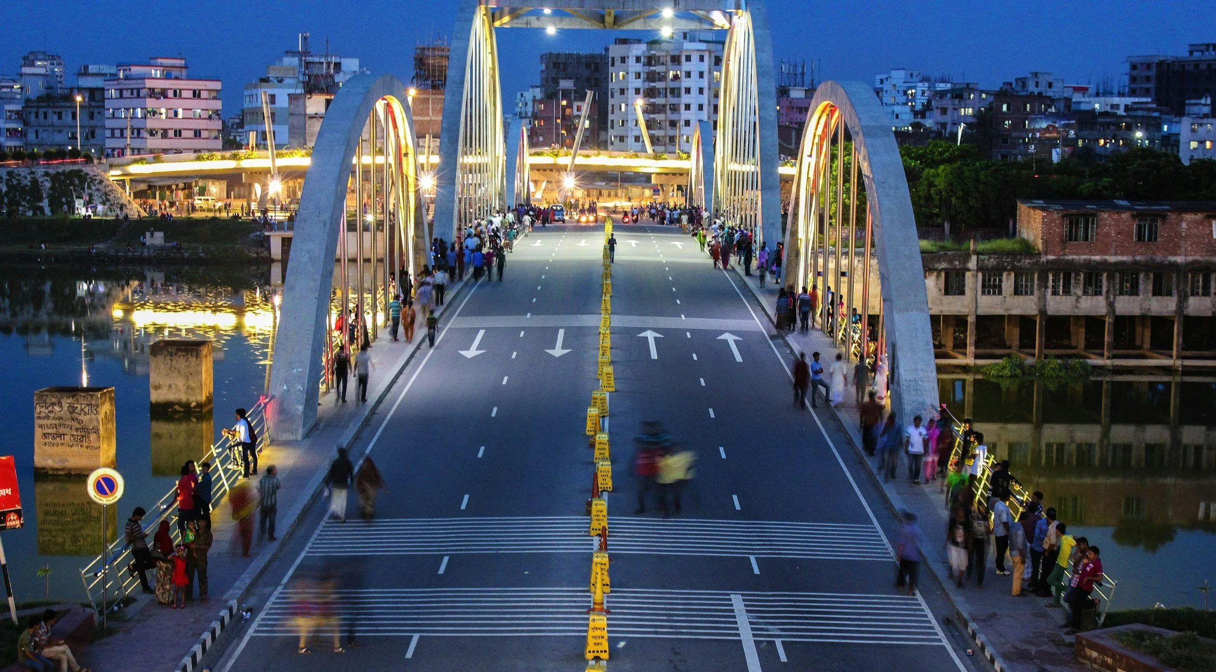 A city bridge with lights at dusk, with people walking on sidewalks on both sides and a blurred bicycle crossing. The bridge has large white arches and surrounding buildings are visible, some under construction.