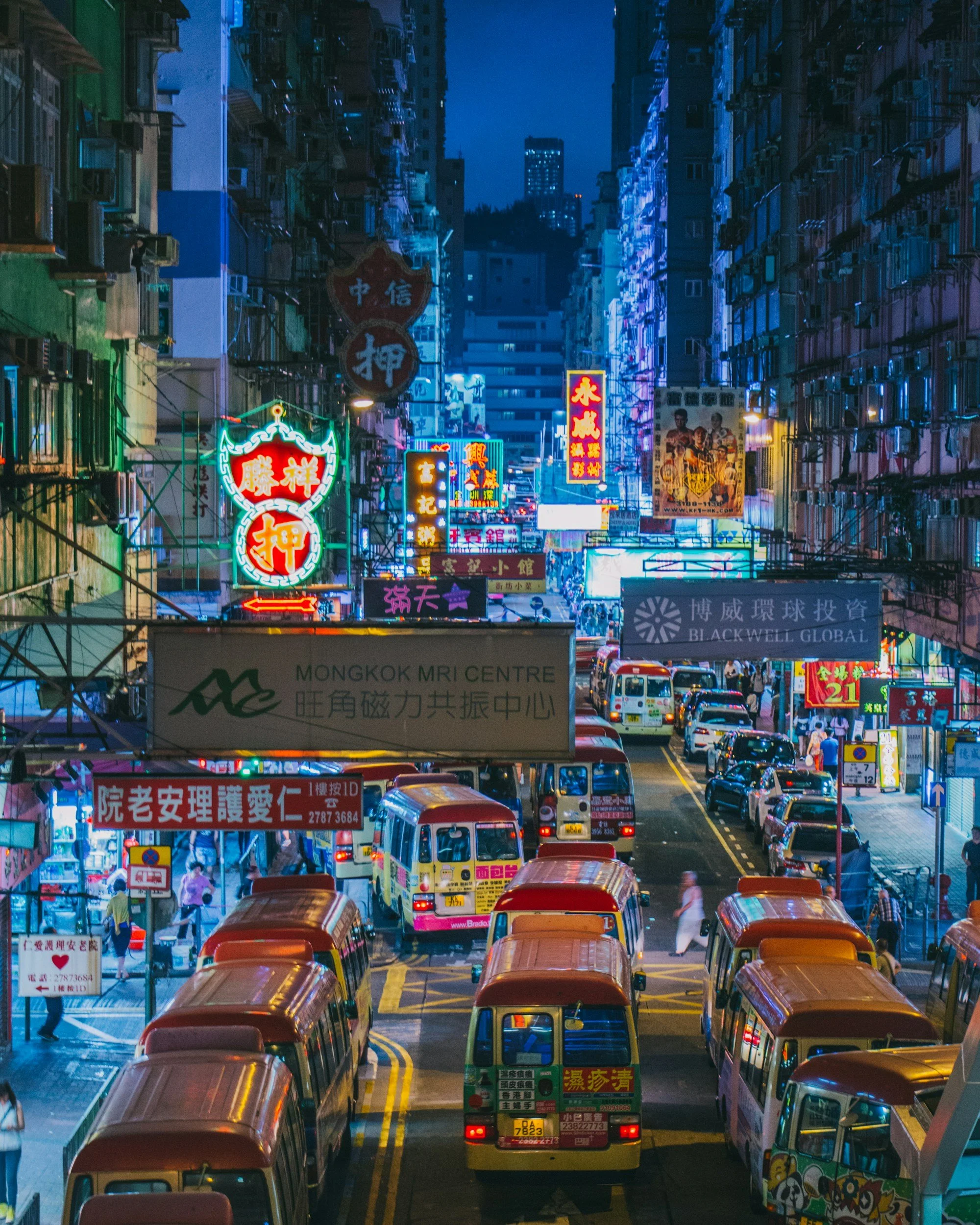 Nighttime street scene in a densely packed urban area with neon signs in Chinese characters, traffic, and people walking.