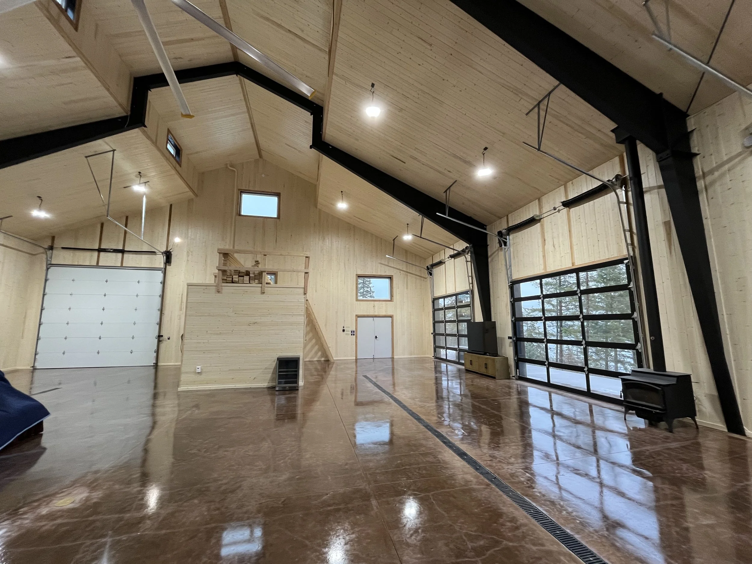 Interior of a spacious, modern garage with wooden walls and ceiling, epoxy-coated floor, large glass garage doors, and minimal furnishings.