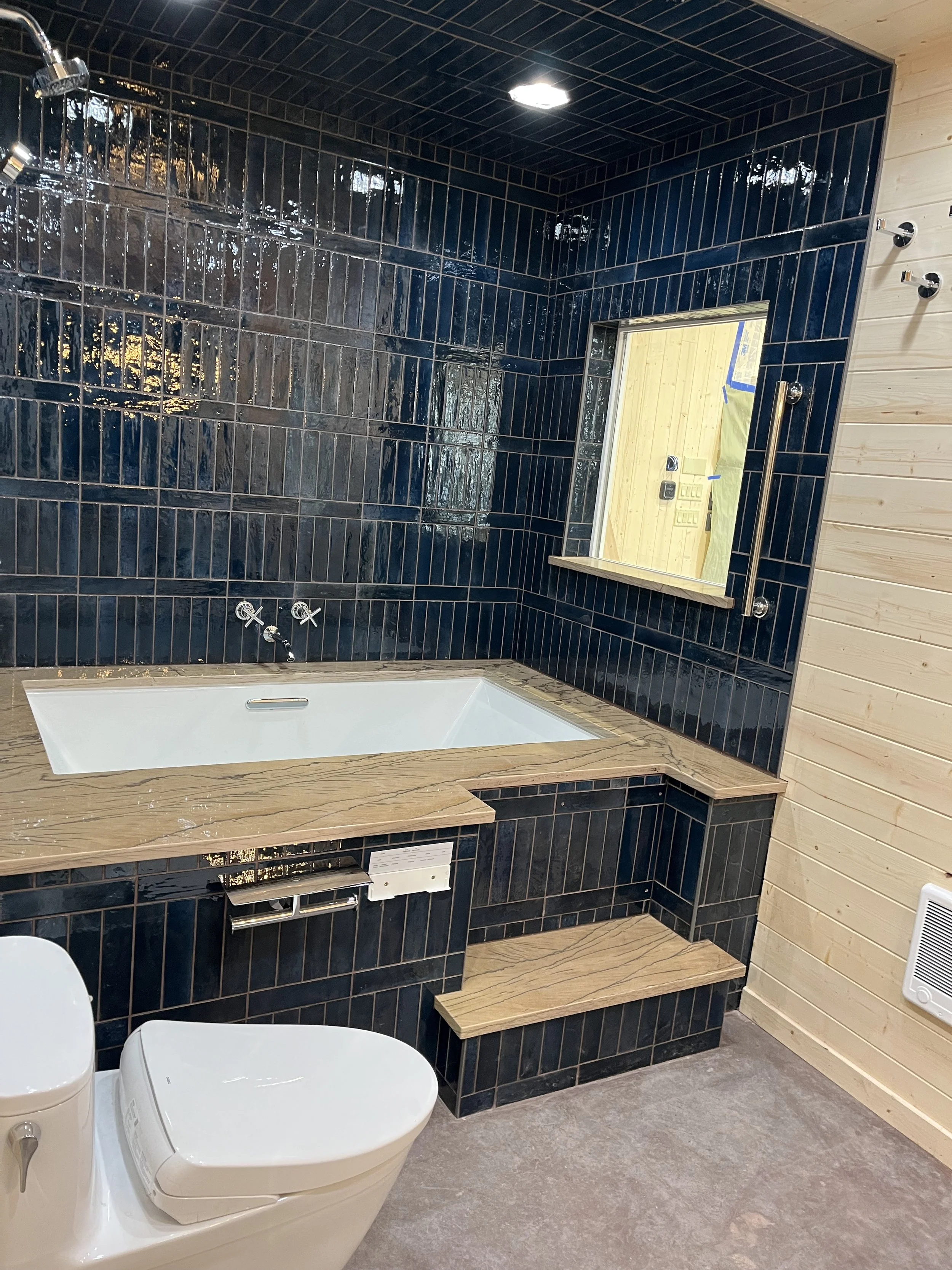 Bathroom with dark blue tiled walls, a built-in bathtub with a wooden surround, a mirror, a toilet, and a wooden-paneled wall.
