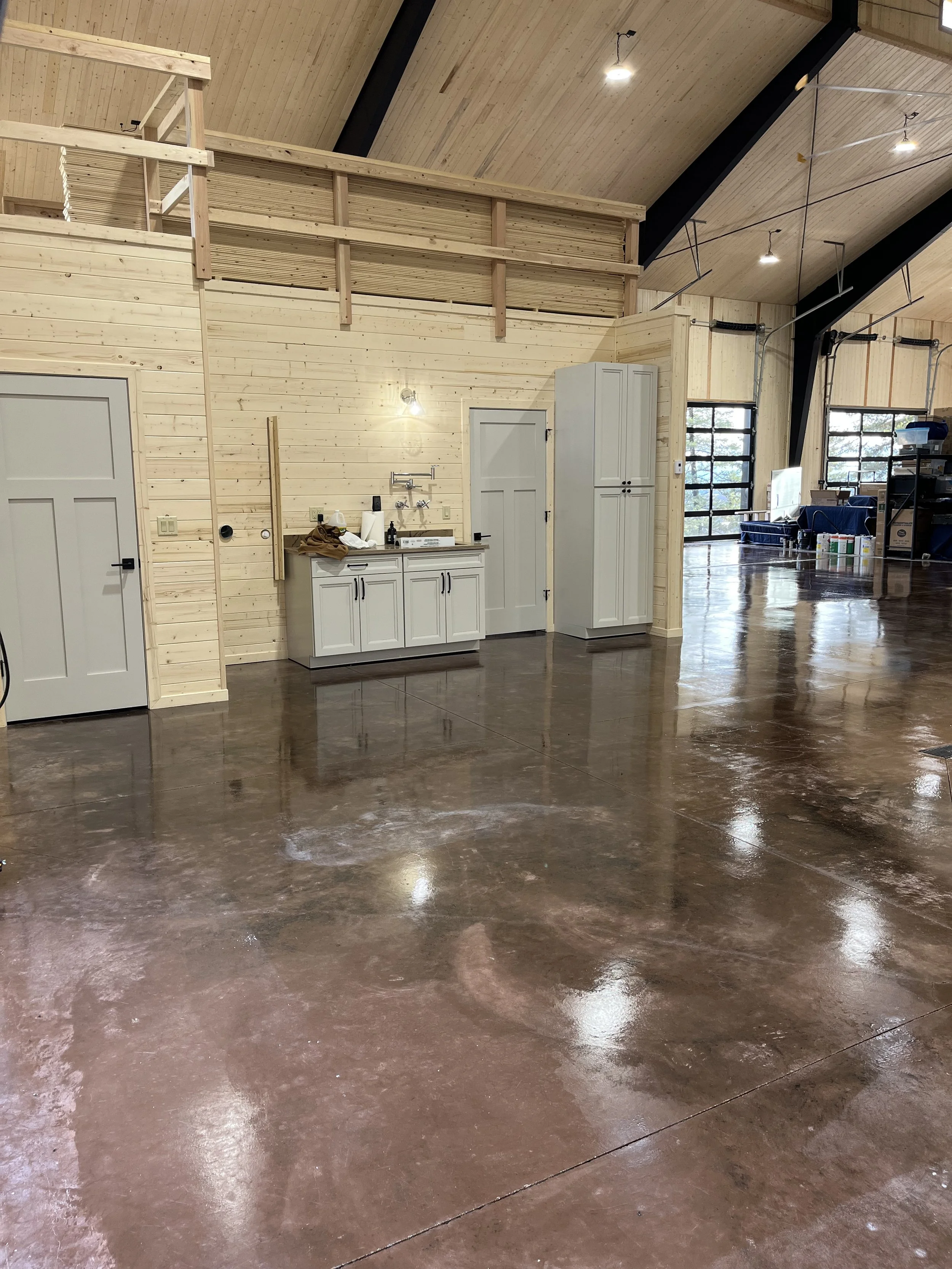 A spacious garage or workshop with polished concrete floors, wooden wall paneling, a small white sink with cabinets, and a large cabinet. There are windows with black frames, ceiling lights, and a loft area with a railing.