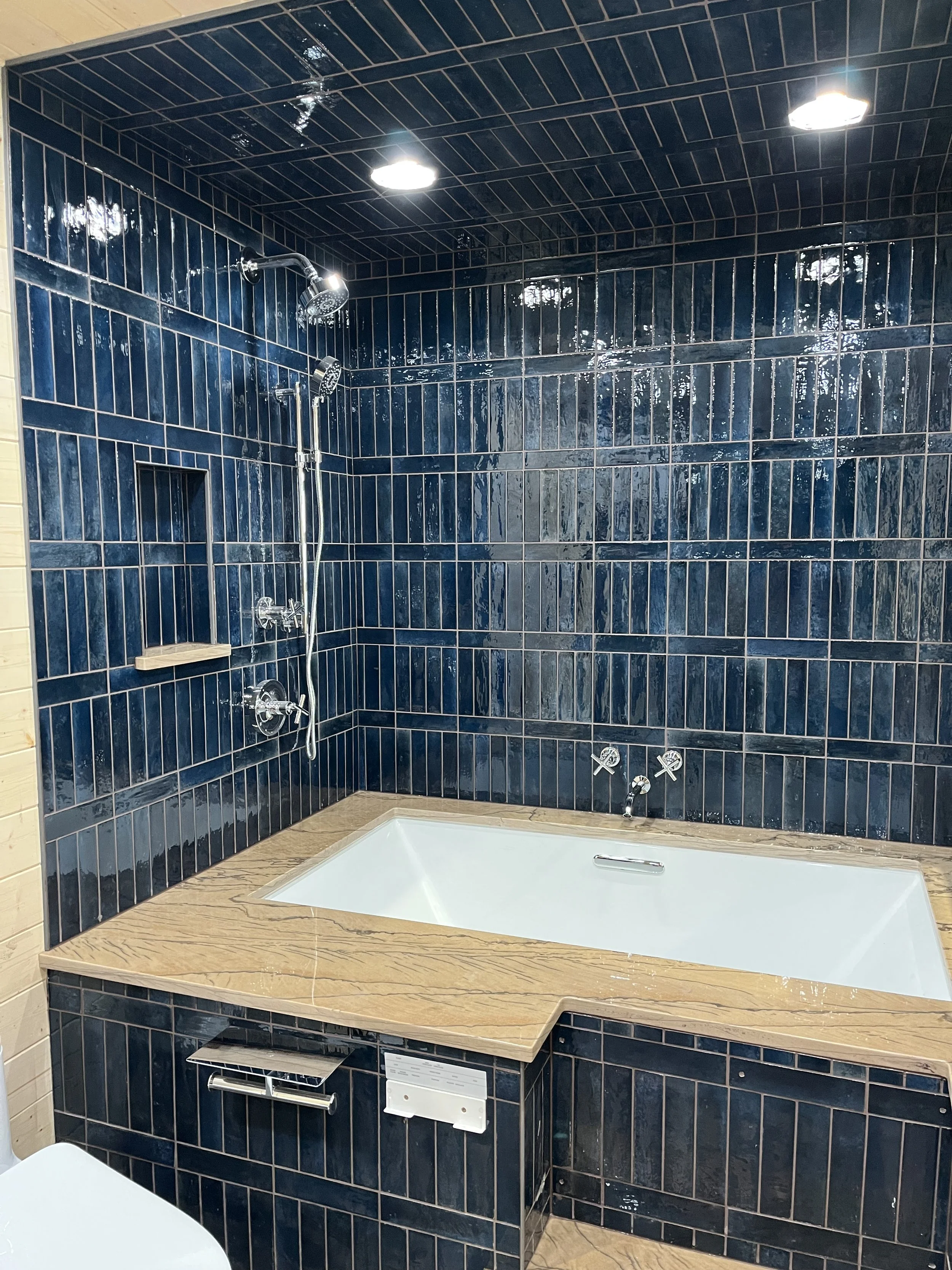 Bathroom with dark blue glossy tiled walls and ceiling, a built-in bathtub with a beige marble surround, a wall-mounted shower system, and overhead lighting.