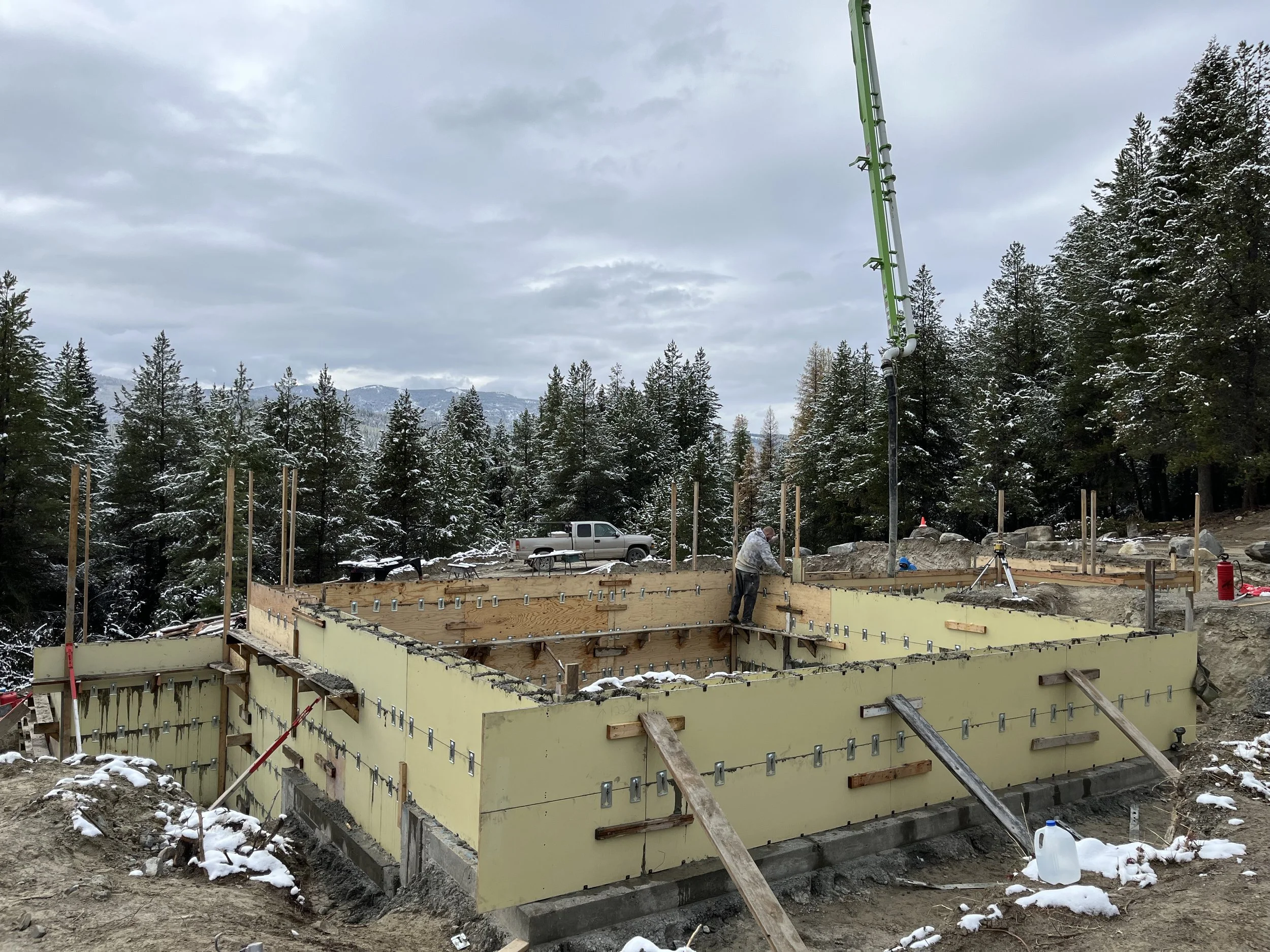 Construction site with foundation walls being built, surrounded by snow and trees, with a worker working on the foundation