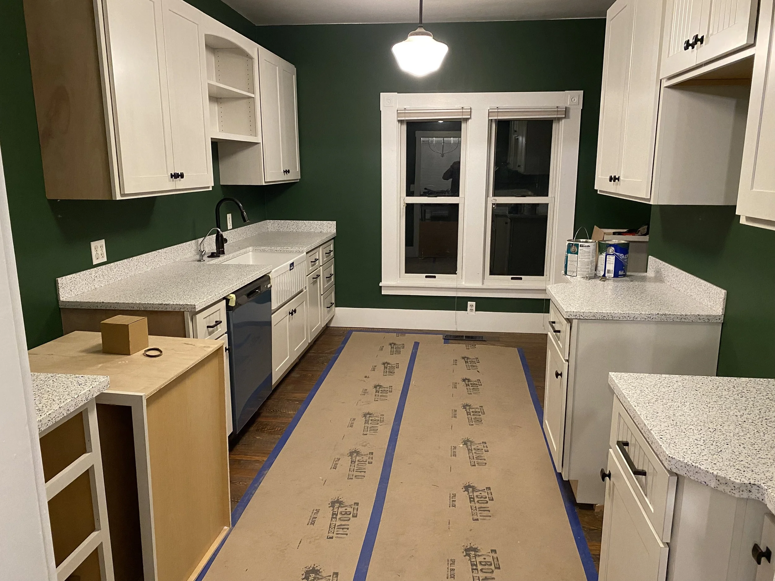 Kitchen with white cabinets, green walls, hardwood flooring, and a window with a white frame. The countertops are speckled gray. There are some paint cans on the right counter, and a cardboard box on the left counter. A ceiling light is turned on, an
