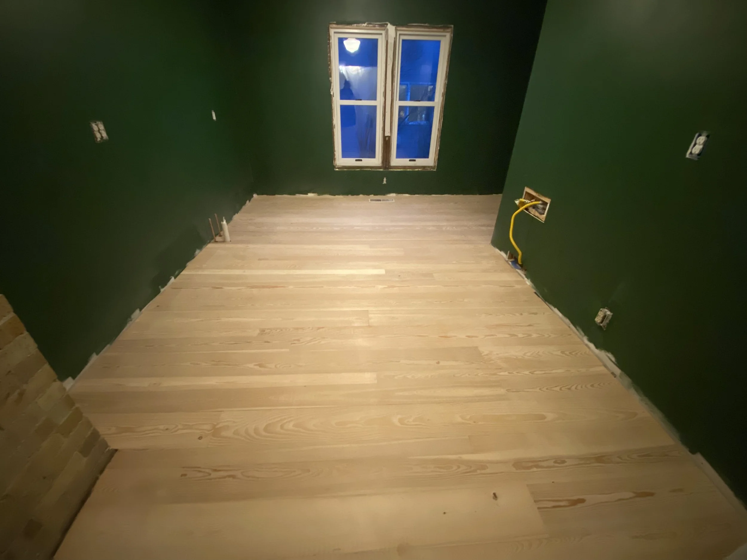 An empty room with green walls and light-colored hardwood flooring, with a window on the far wall and electrical outlets and wiring visible during construction or renovation.