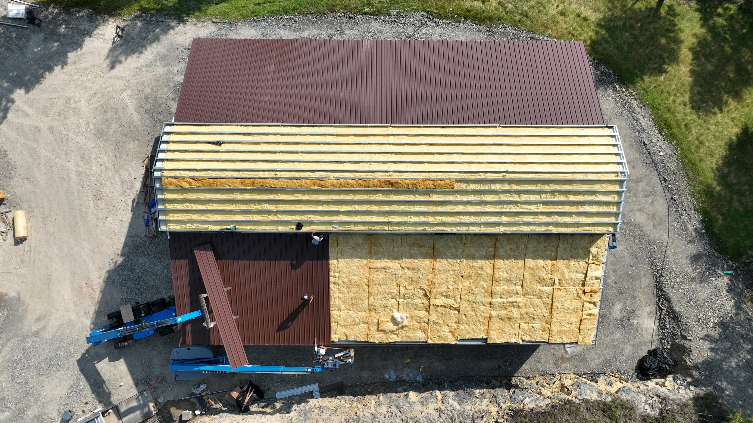 Aerial view of a building under construction with a roof being installed. Part of the roof has brown metal panels, while another part is being insulated with yellow spray foam.