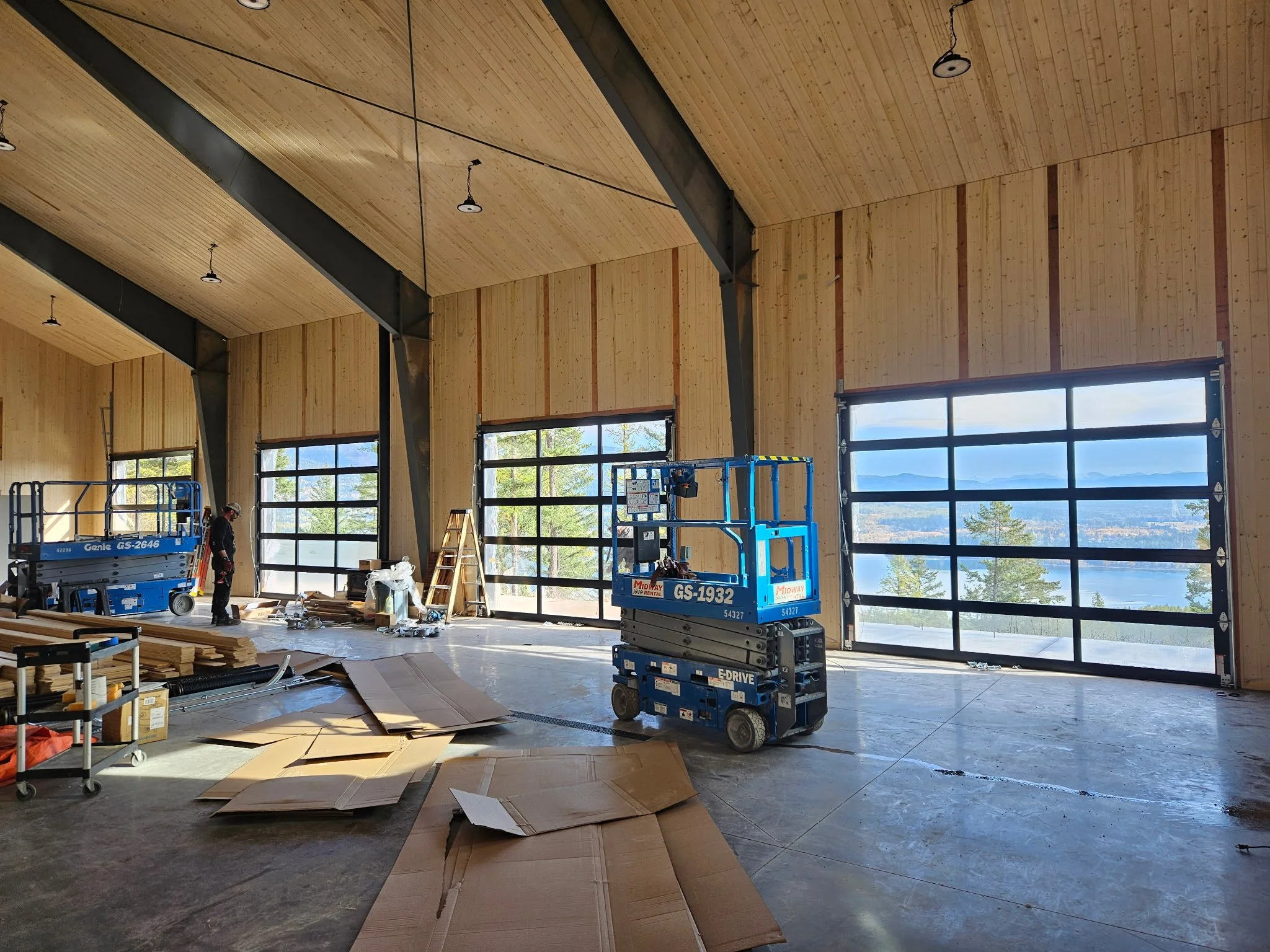 Interior of a building under construction with large windows, wooden walls and ceiling, construction equipment like a lift and ladder, and construction materials on the floor.