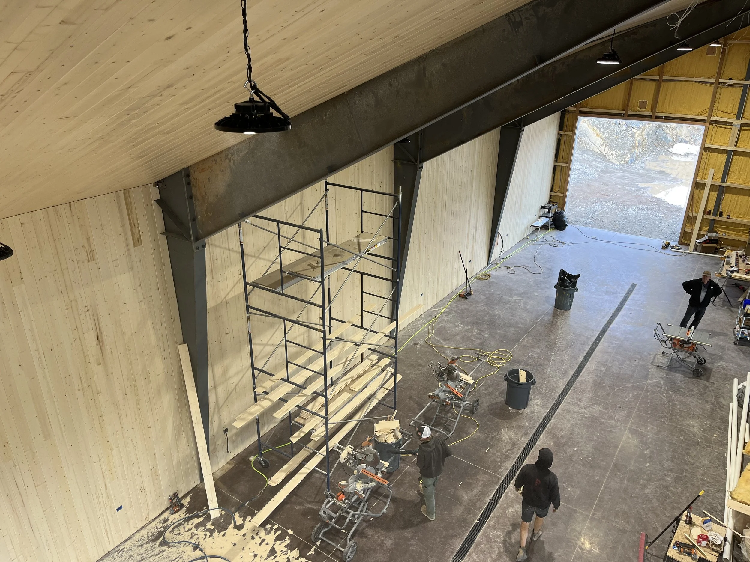 Interior of a large building under construction with high ceilings, scaffolding, and construction workers. There are tools, equipment, and wood framing materials visible.