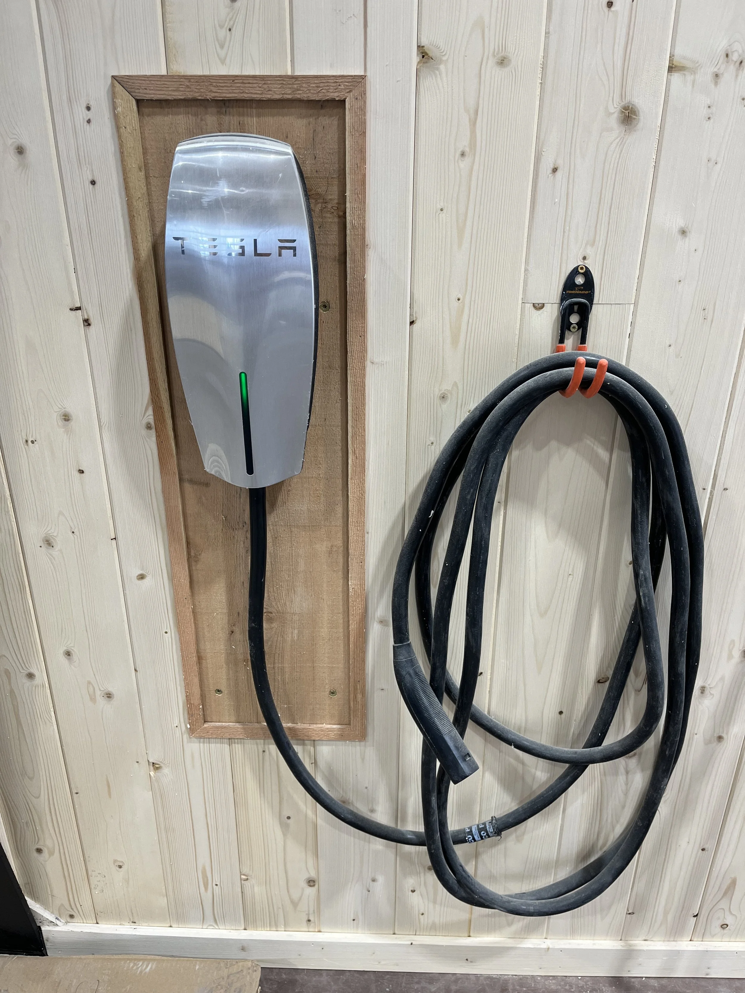 Tesla wall charger mounted on a wooden panel next to a coiled electric vehicle charging cable hanging on a hook on a wood-paneled wall.