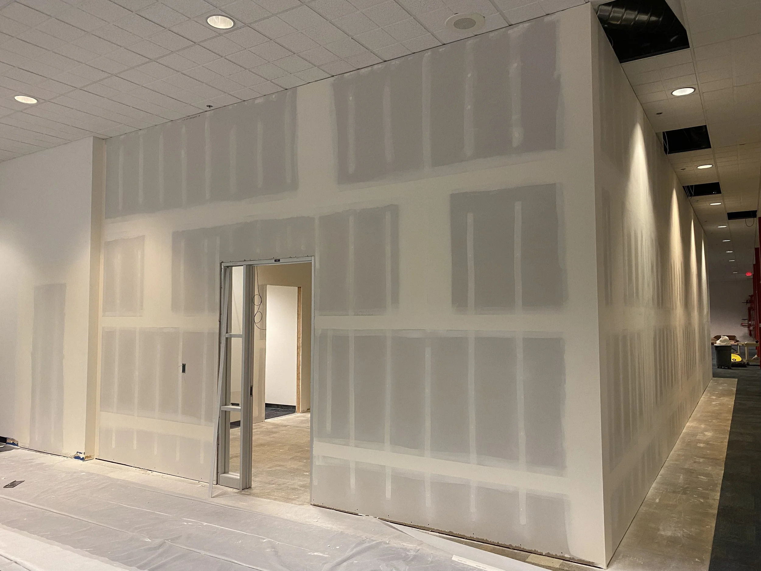 Interior under construction with drywall partially installed on walls and ceiling, with an open doorway leading to another room, in a commercial or office building.