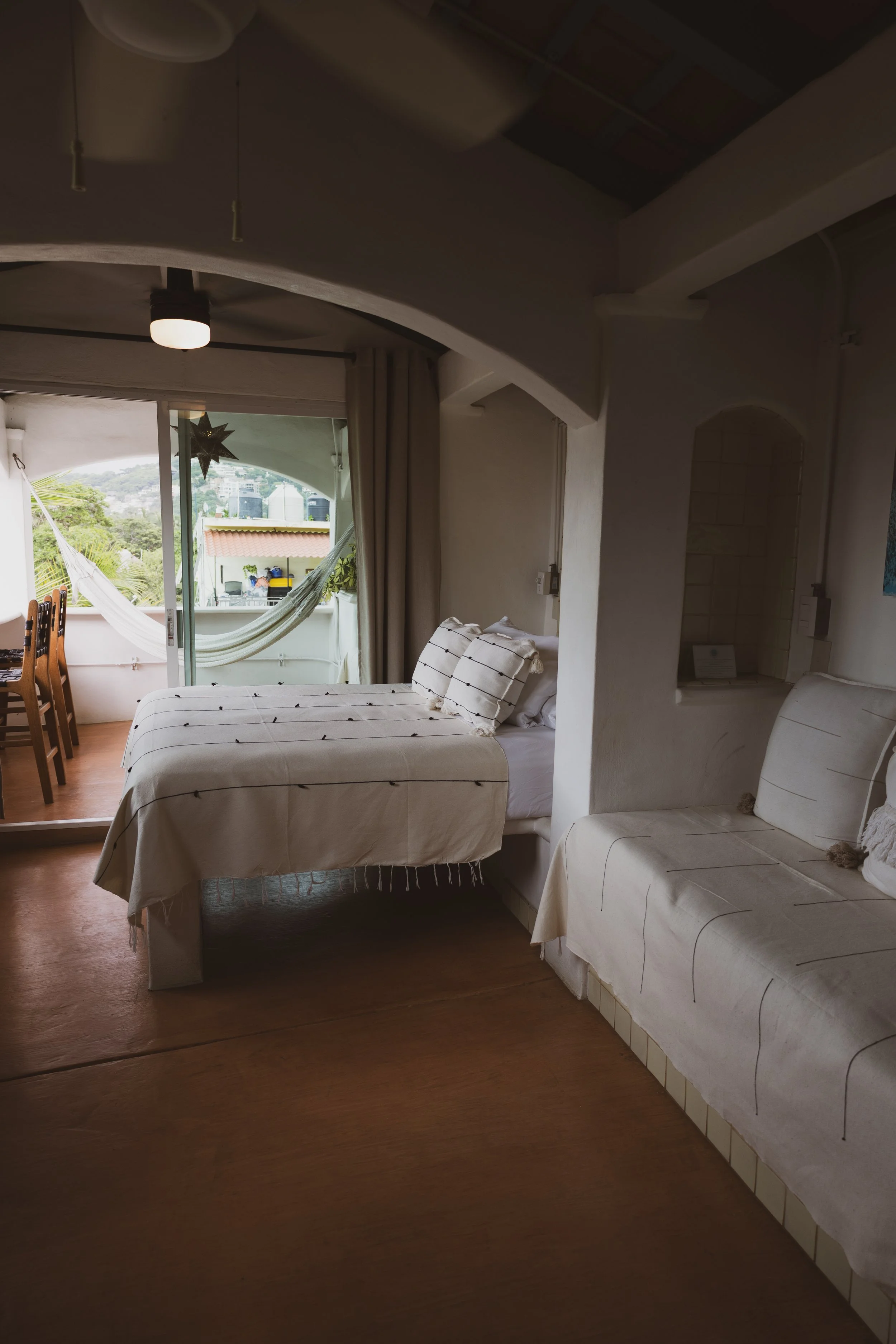 A bedroom with a twin bed and a couch, featuring white linens with black stripes. A sliding glass door leading to a balcony with a hammock and outdoor furniture, views of greenery and nearby buildings.