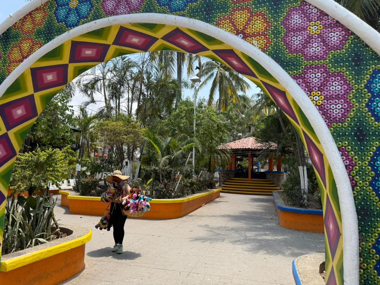 A person walking through a colorful archway in a tropical garden, carrying plush toys and wearing a large sun hat.