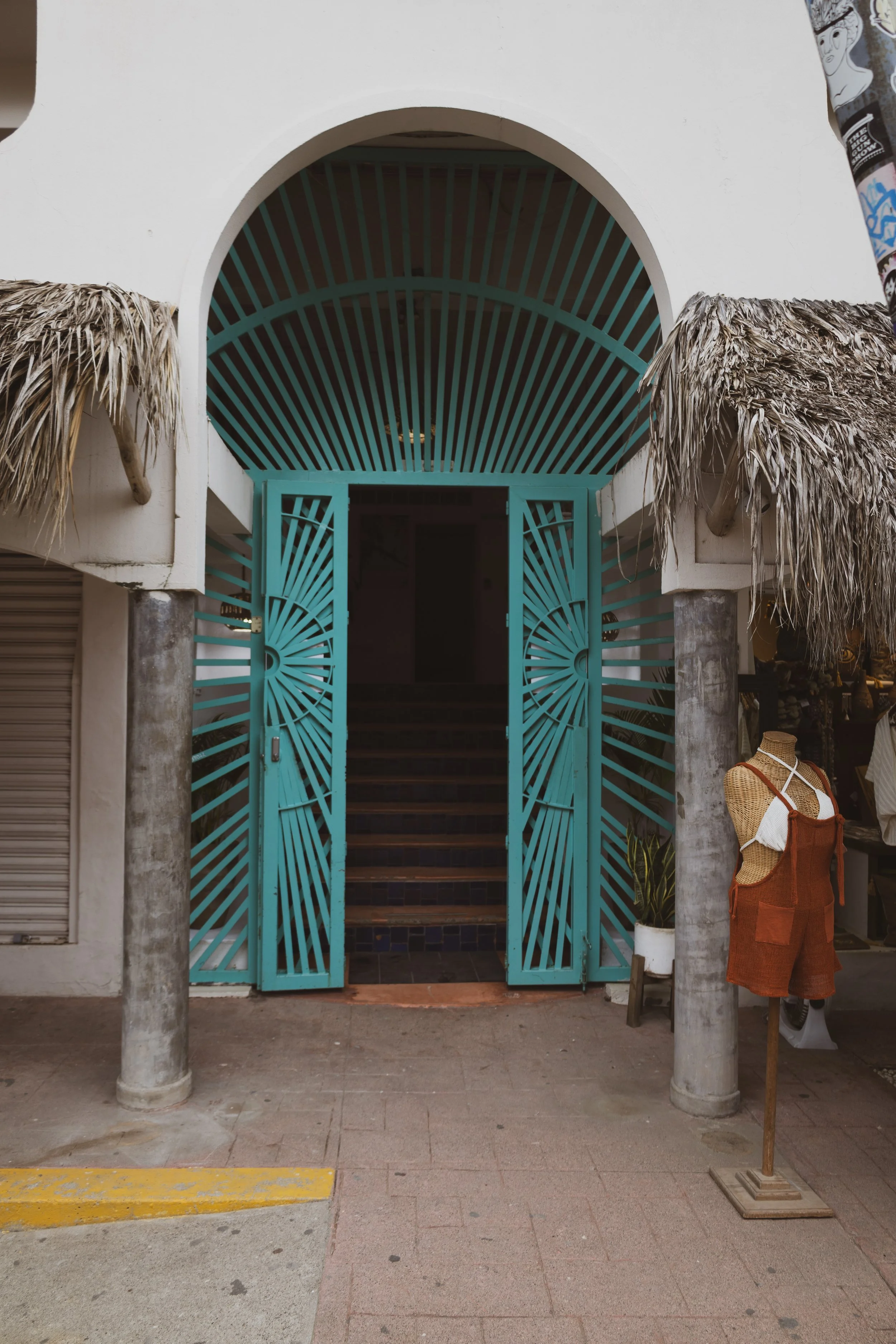 White building entrance with a turquoise gate, arched above with a turquoise and white striped roof, flanked by two concrete columns. There are thatched roof accents on either side of the entrance and a mannequin wearing a crochet top with a brown ba
