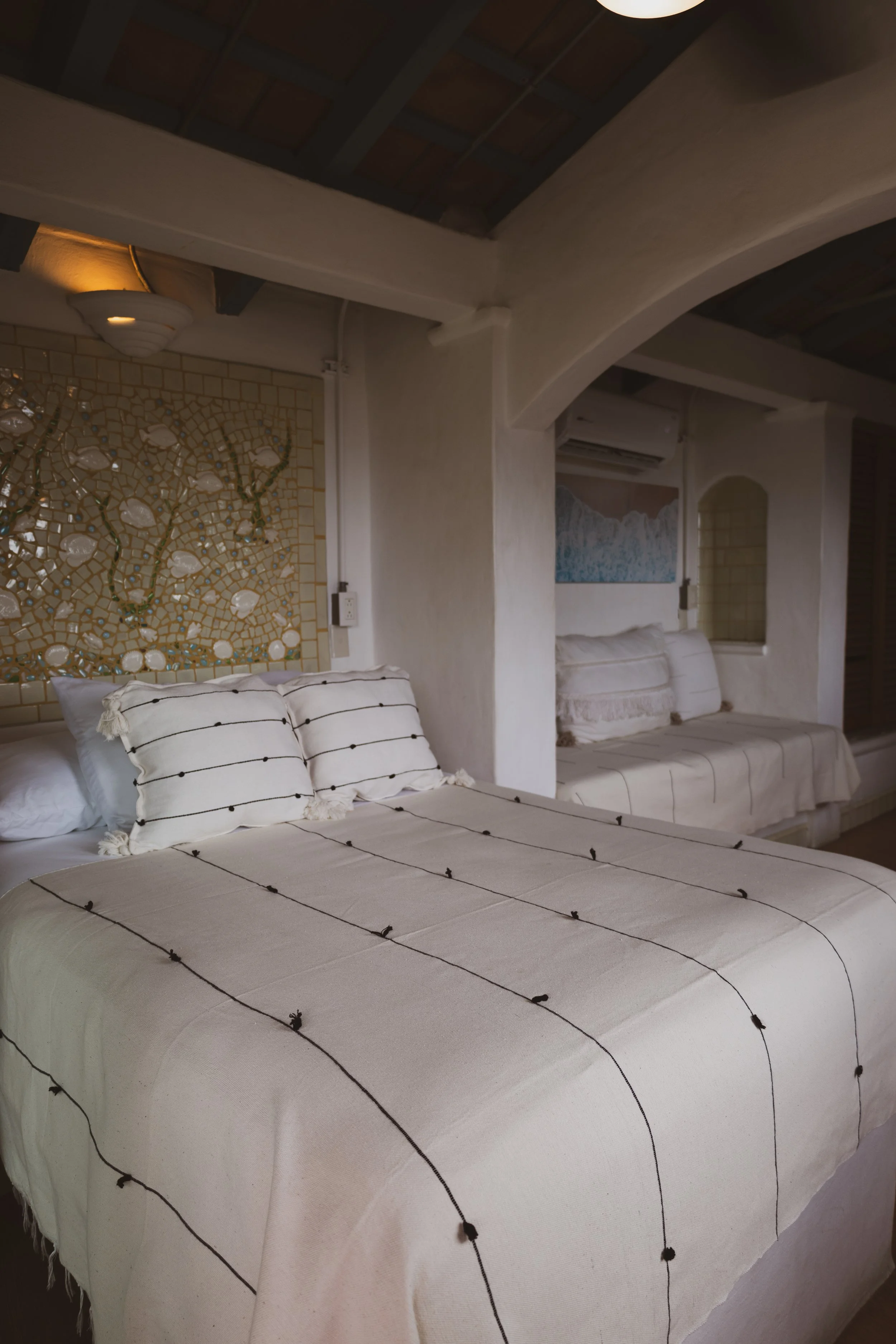 A bedroom with a bed and pillows, decorated with a beige and brown striped bedspread, featuring a mosaic wall decor, and a seating area with cushions in the background.