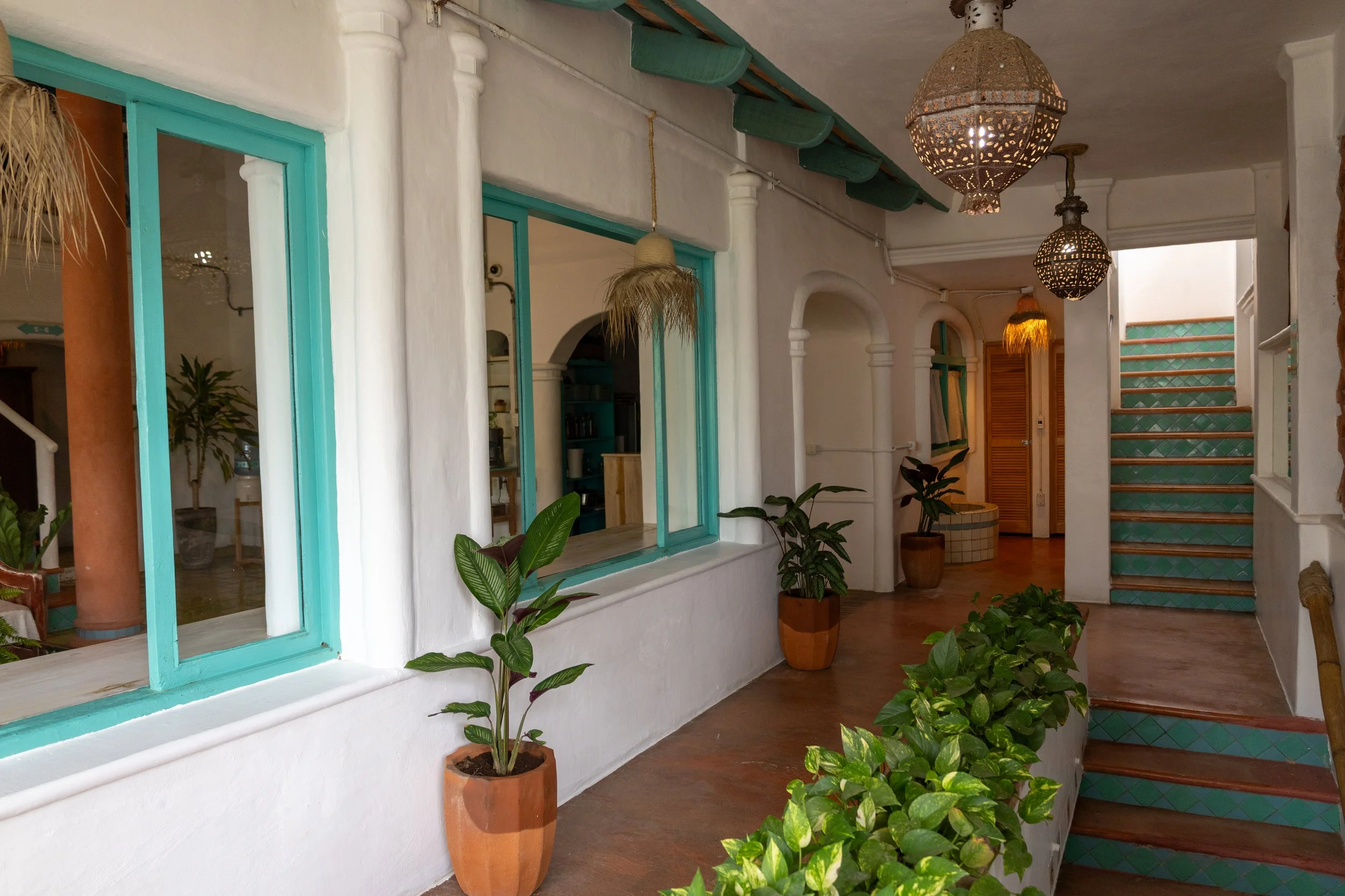 Interior view of a room with white walls, turquoise window frames, green ceiling beams, hanging Moroccan-style lamps, wooden stairs with green tiles, and several potted plants.