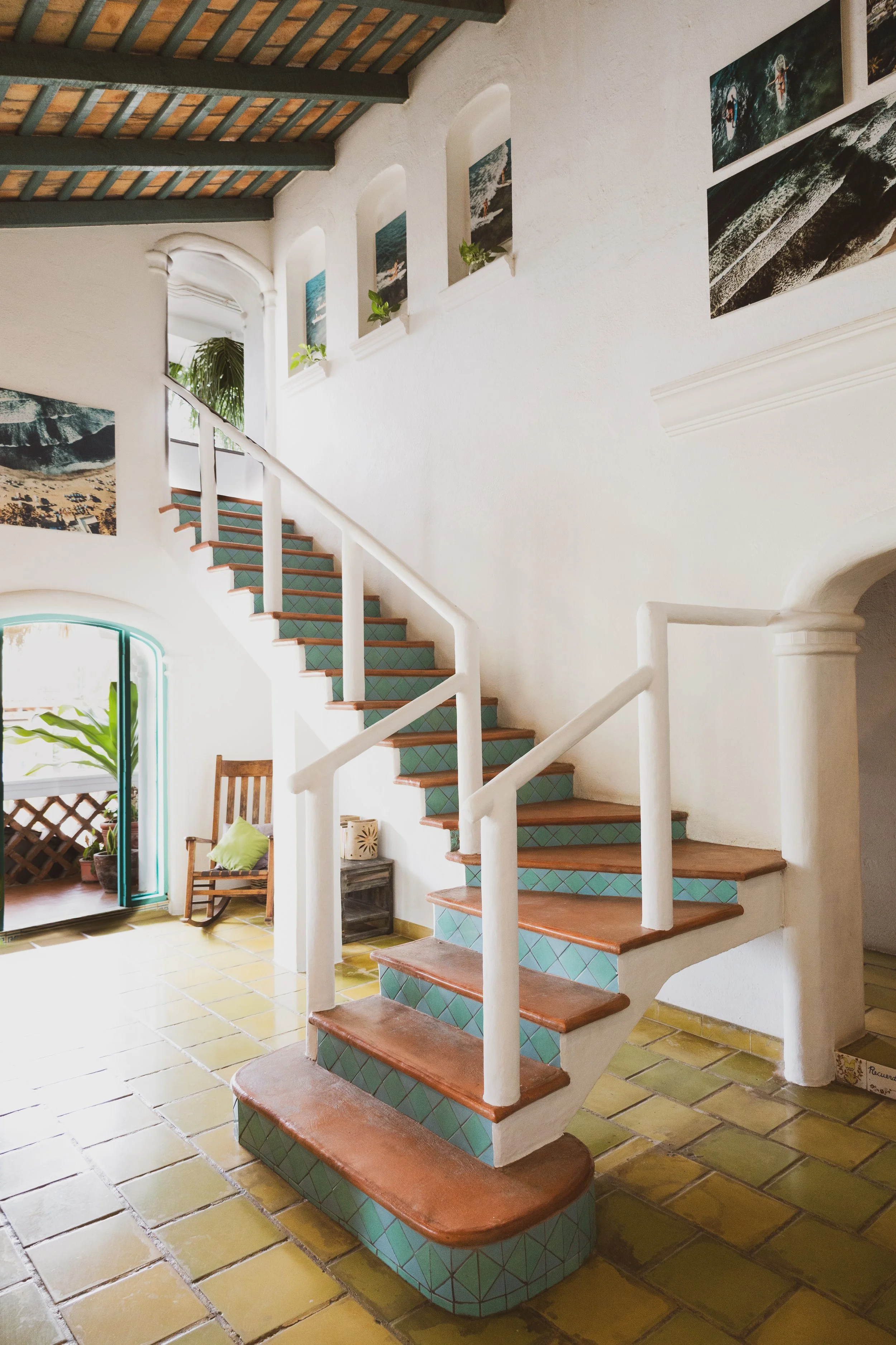 Interior view of a staircase with green tiles and terracotta-colored steps in a bright, open space with white walls, decorative artwork, and potted plants.