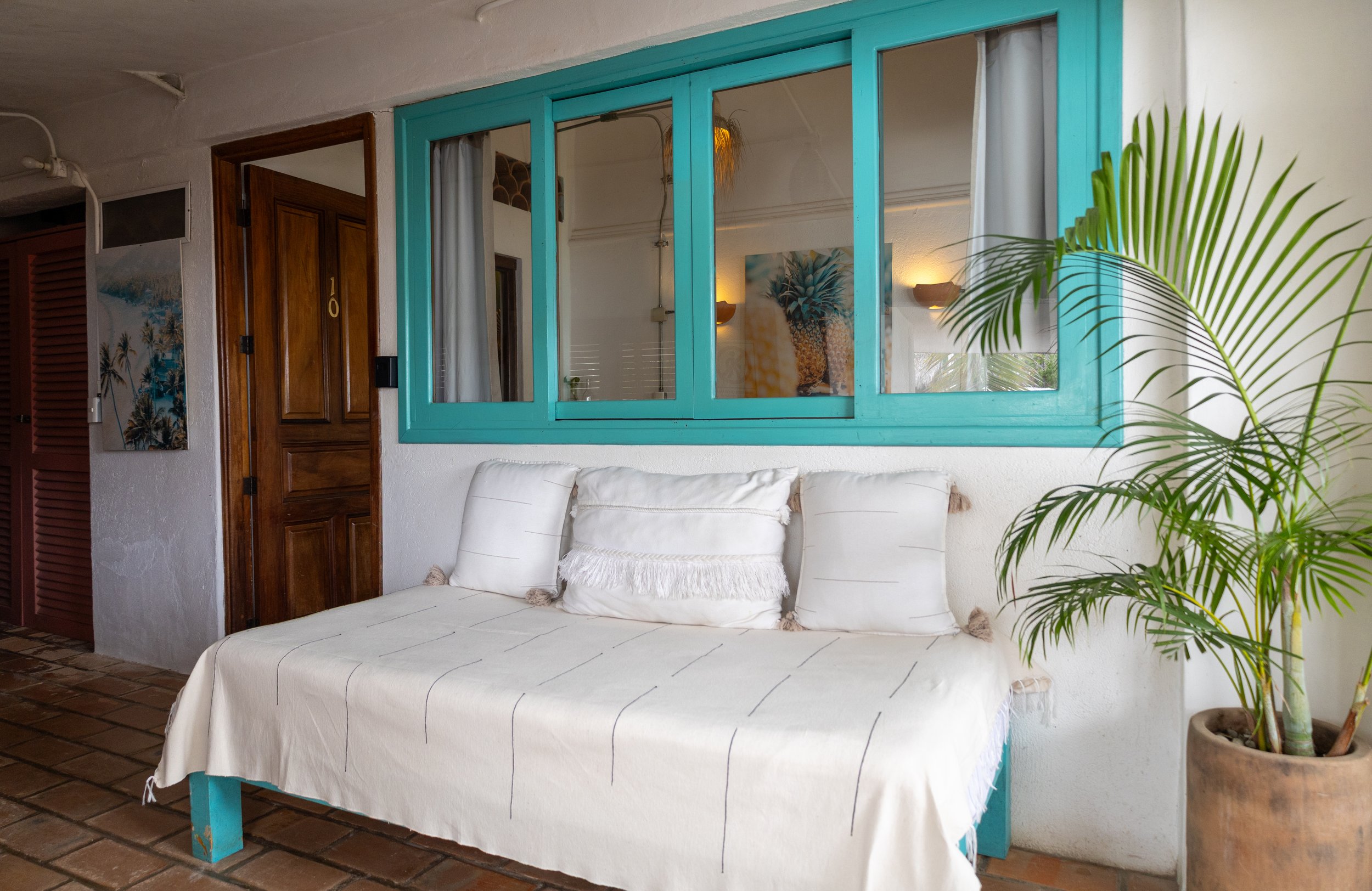 Interior of a room with a white sofa with pillows, a wooden door, a glass window with turquoise frame, also visible are art and a potted palm plant.