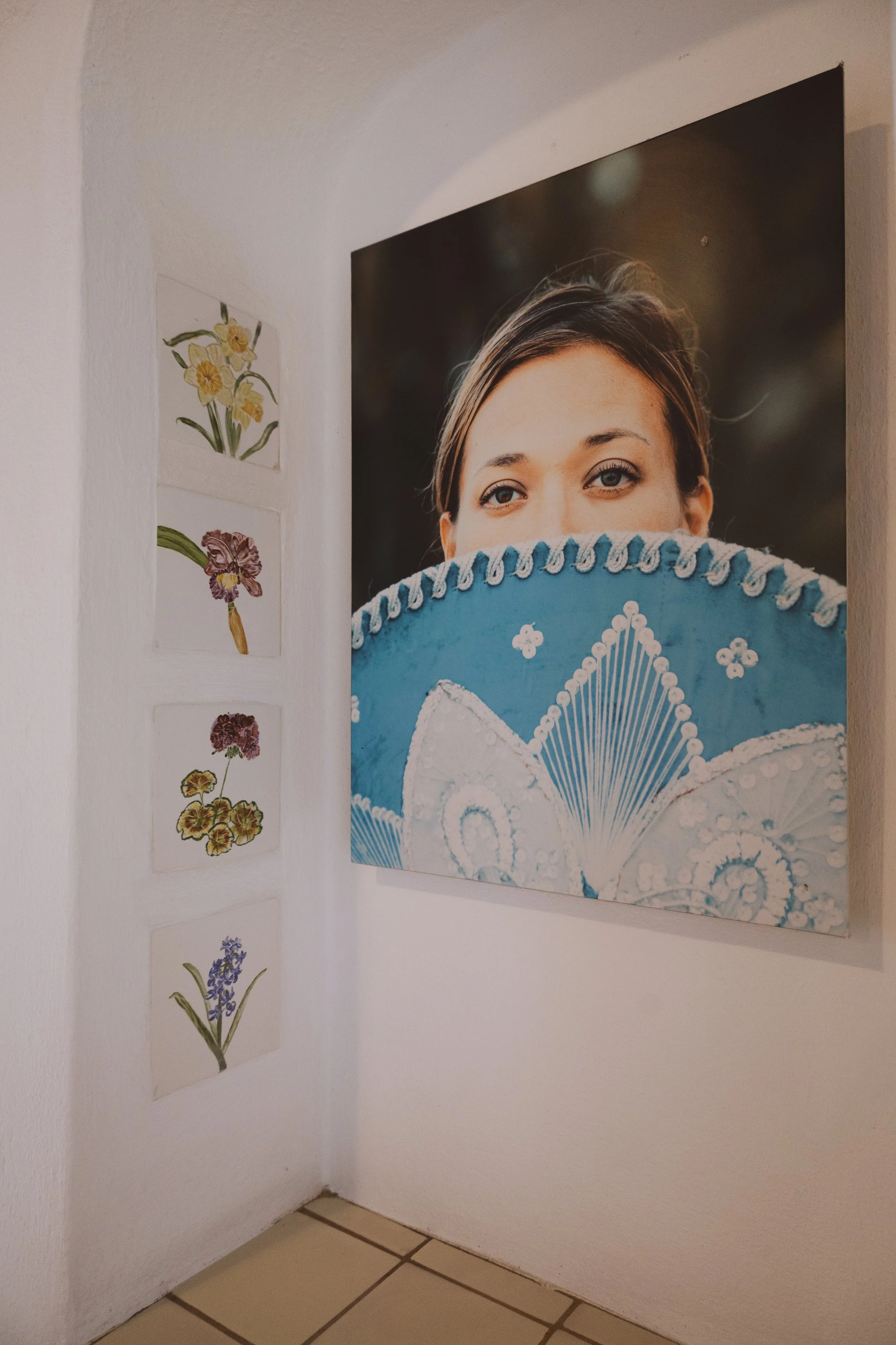 Photo of a corner in a room with white walls, featuring a large portrait of a woman with brown hair holding a decorated blue fan, and a vertical row of five botanical illustrations of flowering plants on the adjacent wall.