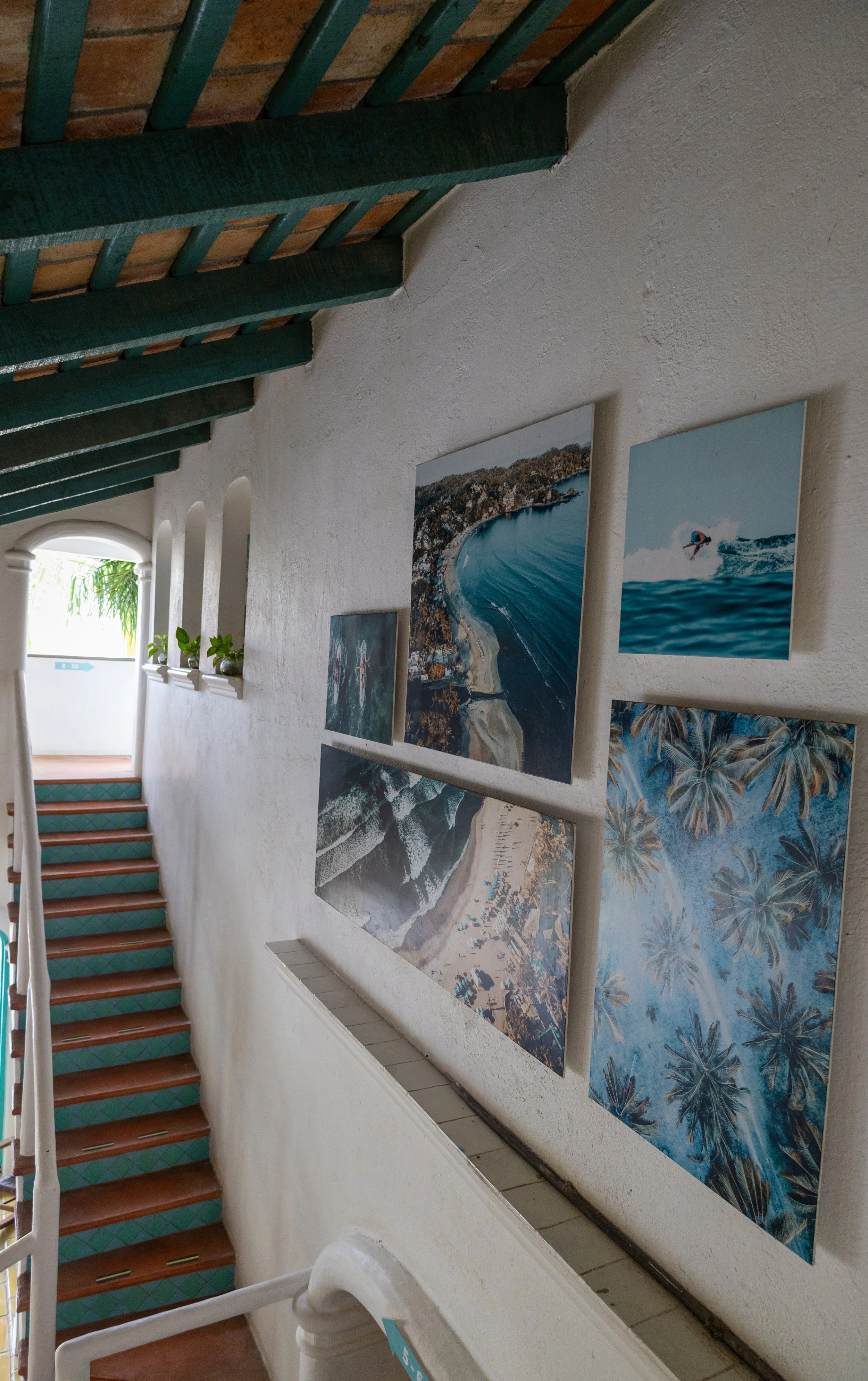 Interior view of a staircase with wooden steps edged in green, beside a white textured wall decorated with large framed photos of beaches, waves, and palm trees.