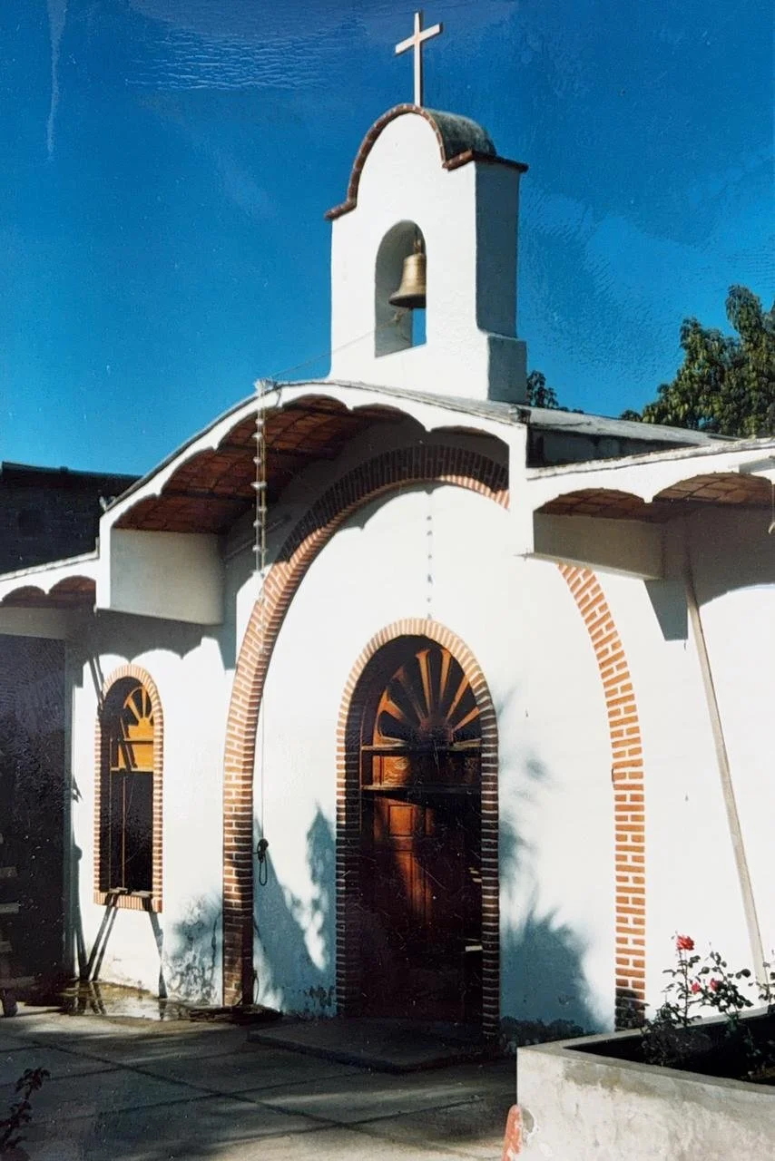 Small white church with a bell tower topped with a cross, arched wooden door, and a flower bed in front, under a clear blue sky.