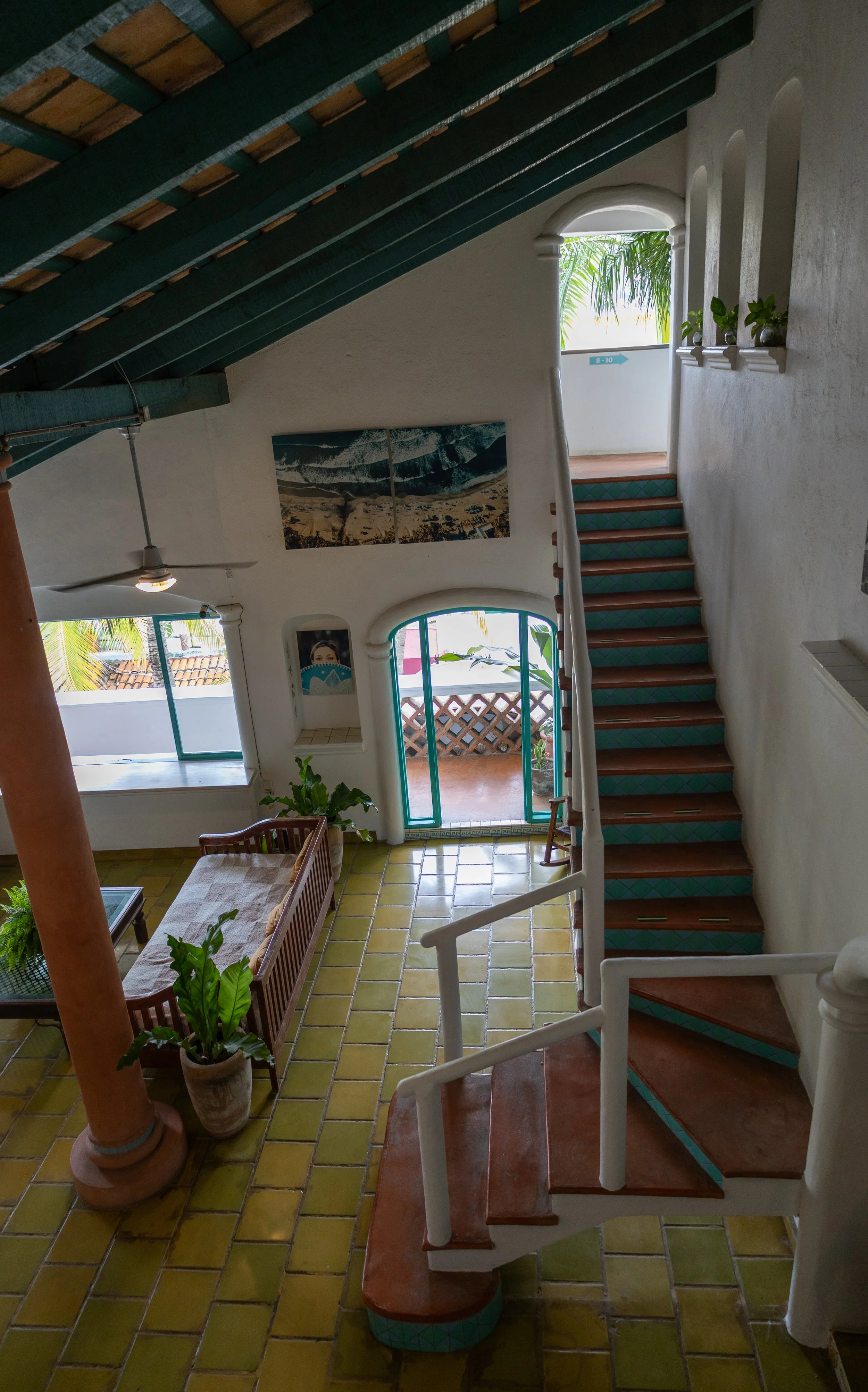 Interior view of a two-story house with yellow tiled flooring, a wooden staircase with green tiles on the risers, a porch with potted plants, large windows, and colorful artwork on the walls.