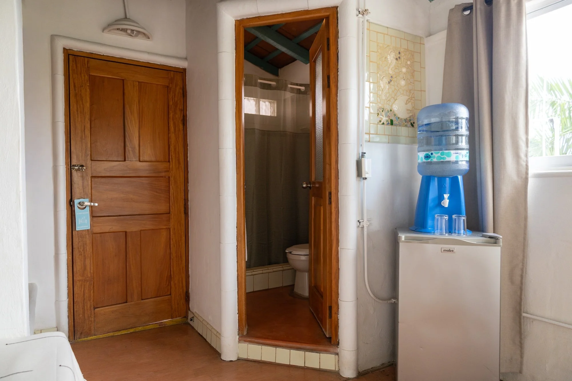 Interior of a room with a wooden door, a small bathroom with a toilet, a water dispenser, and a window with curtains.