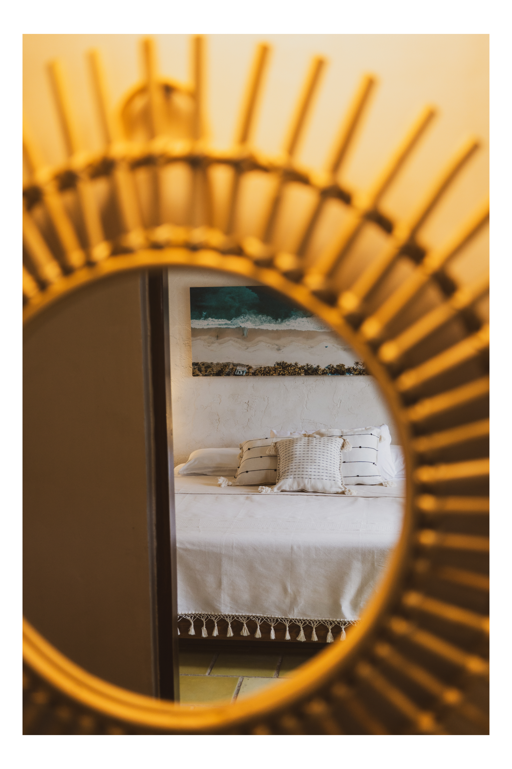 Bedroom with a bed covered in a white bedspread, decorated with striped pillows and a decorative fringe. A landscape painting of a beach hangs on the wall behind the bed. The photo is taken through a round mirror with a woven, golden frame.