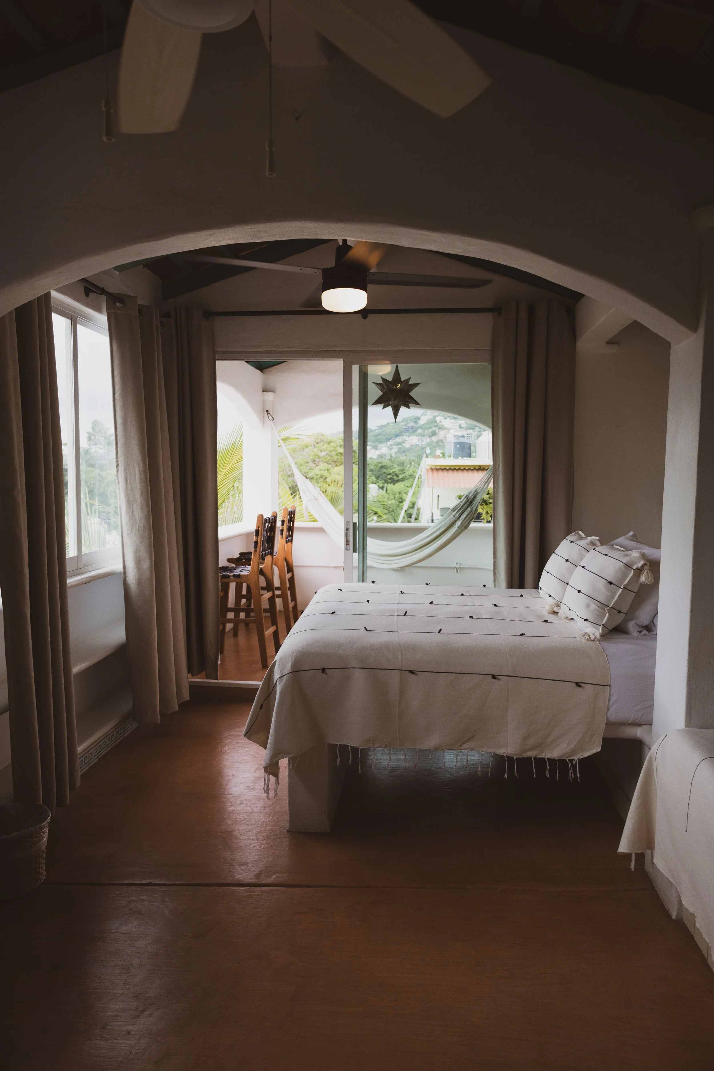 A cozy bedroom with a bed covered in beige linens, a window with beige curtains, and a balcony with outdoor chairs and a hammock.
