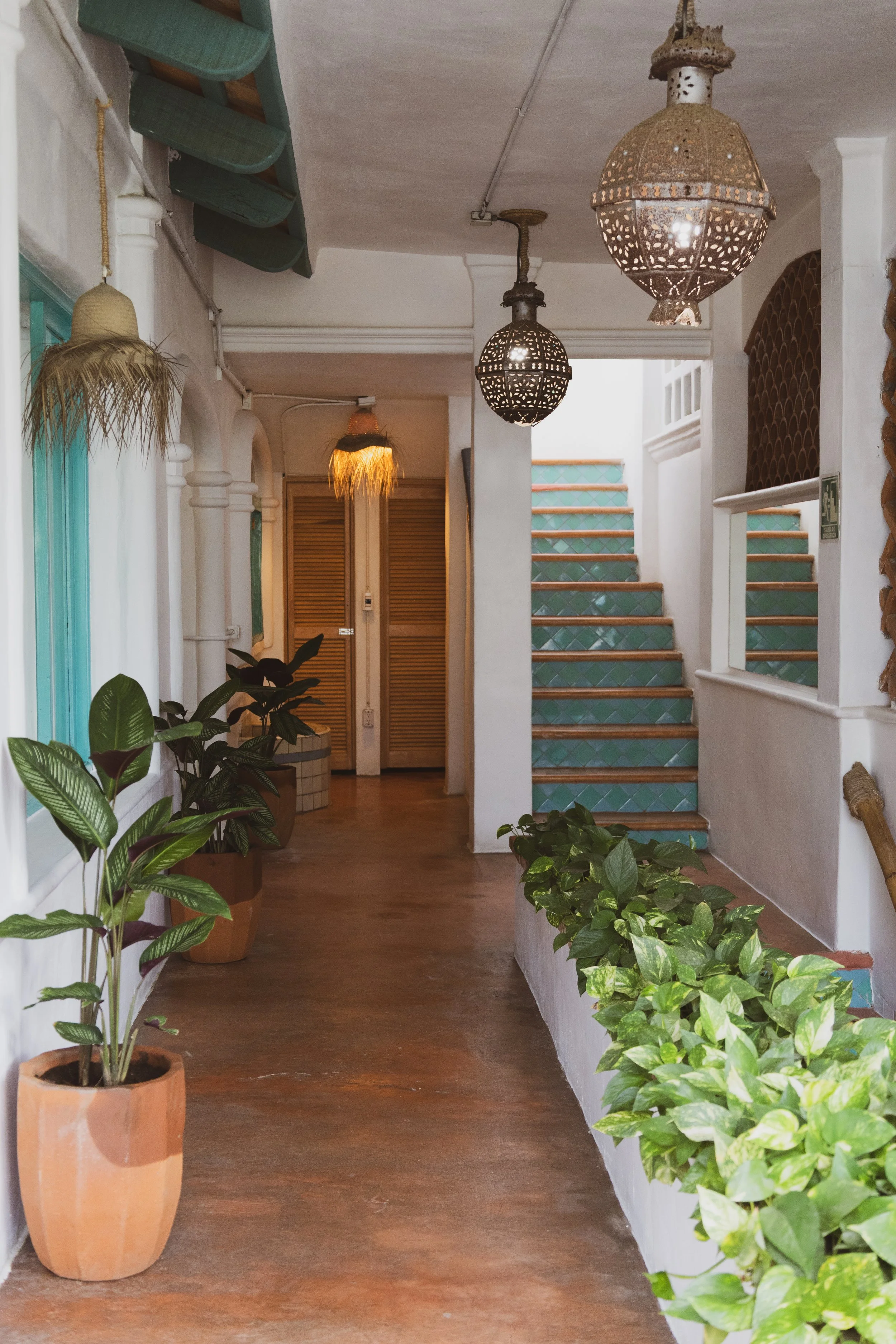Interior shot of a cozy, Mediterranean-style hallway with potted plants, decorative hanging lamps, and green tile stairs with wooden edges.
