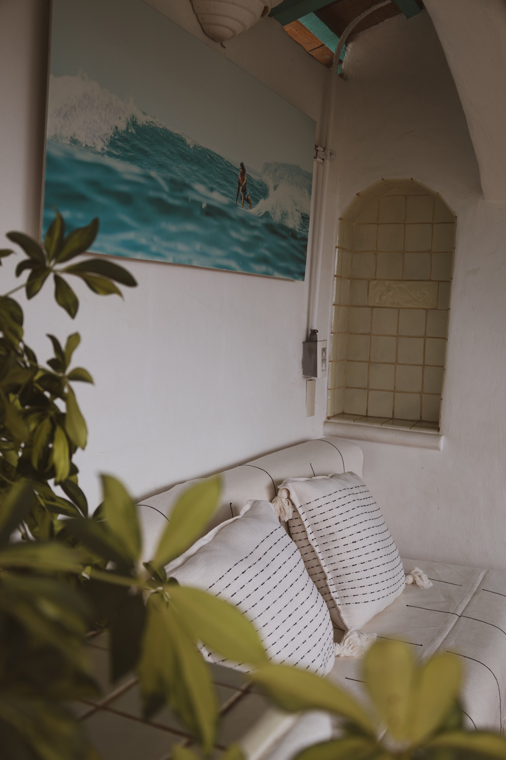 A cozy corner of a room with a white couch decorated with black-striped pillows. A painting of a surfer riding a wave hangs on the wall above the couch. To the right, there's a small arched window with tiles inside and a white wall.