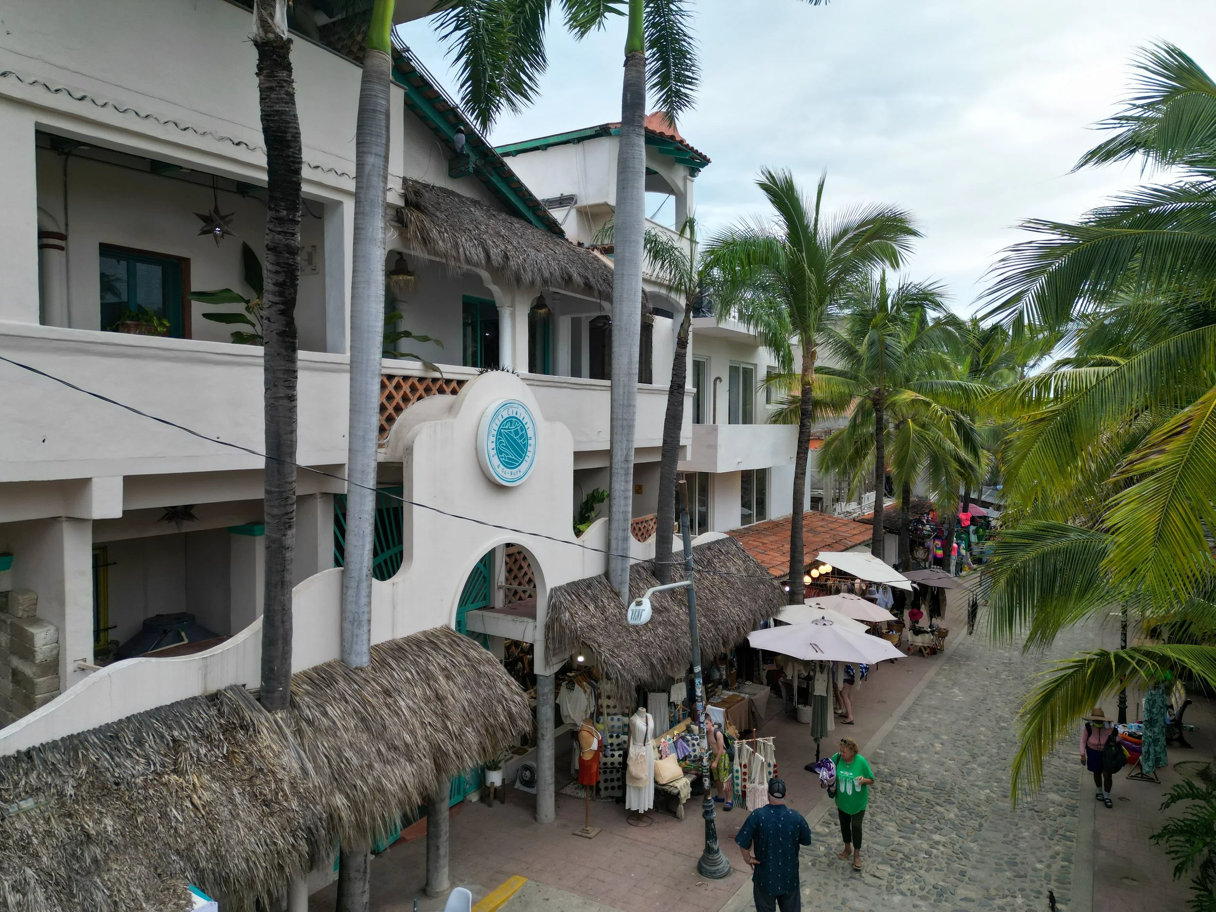 A busy outdoor shopping street with white buildings, straw thatched roofs, tall palm trees, and market stalls with clothes and souvenirs. People are walking around shopping and enjoying the scene.