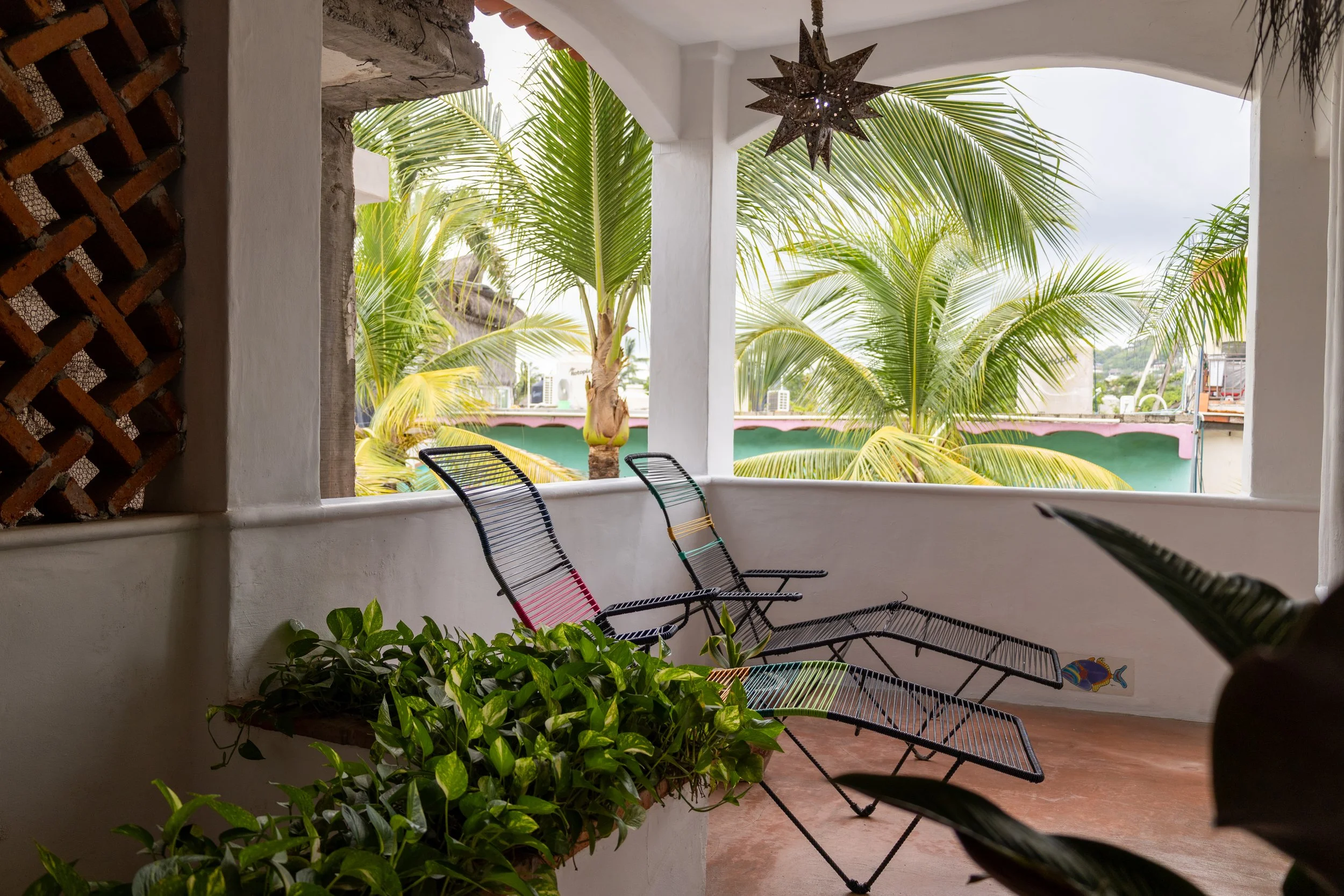 View from a balcony with three black metal chairs, green potted plants, and a white wall. The background features palm trees and a cloudy sky.