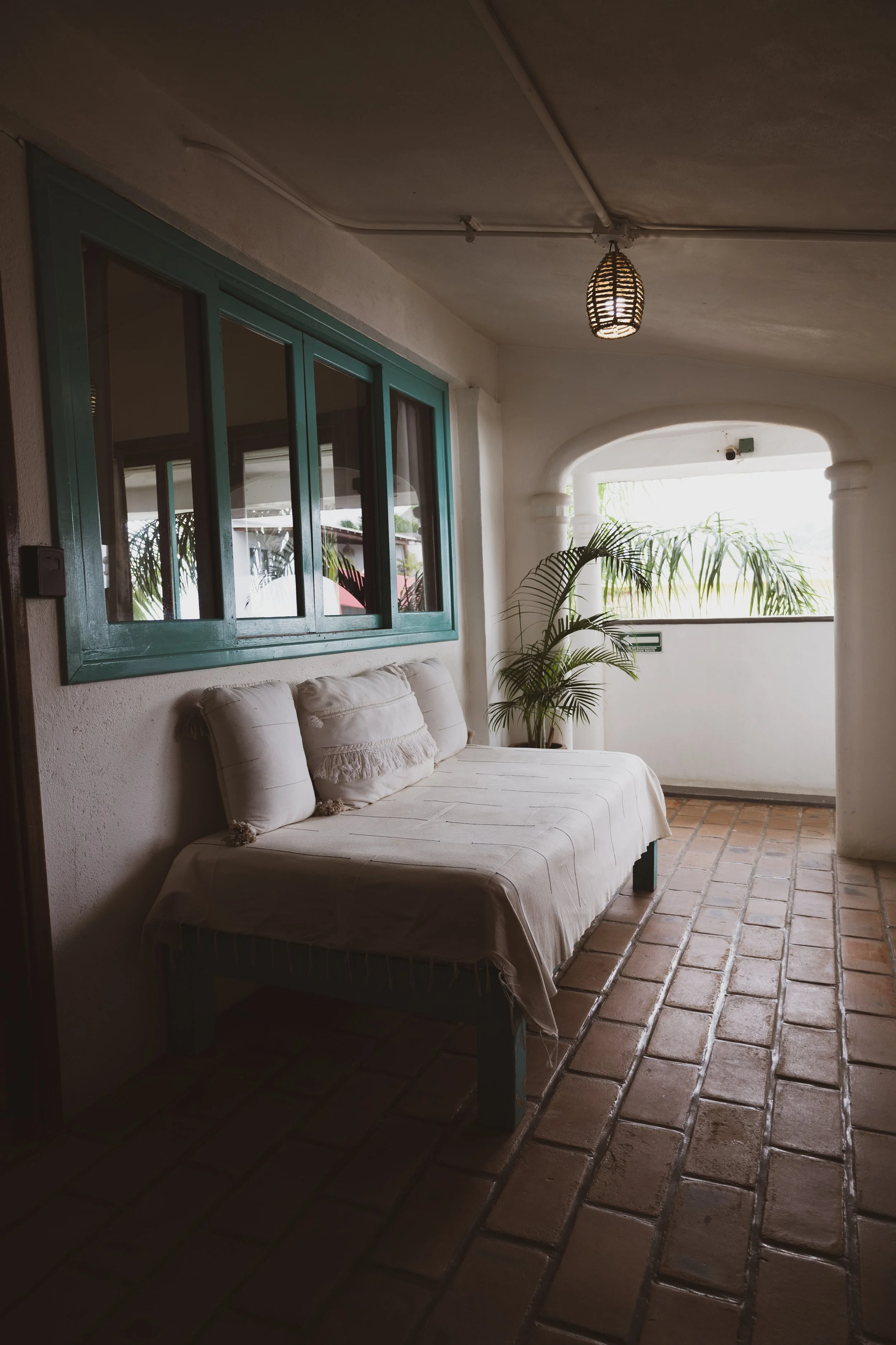 A cozy outdoor patio space with a white cushioned bench, beige pillows, a potted plant, a teal window, and a hanging wicker lantern on a ceiling, with sunlight coming in from an arched opening.