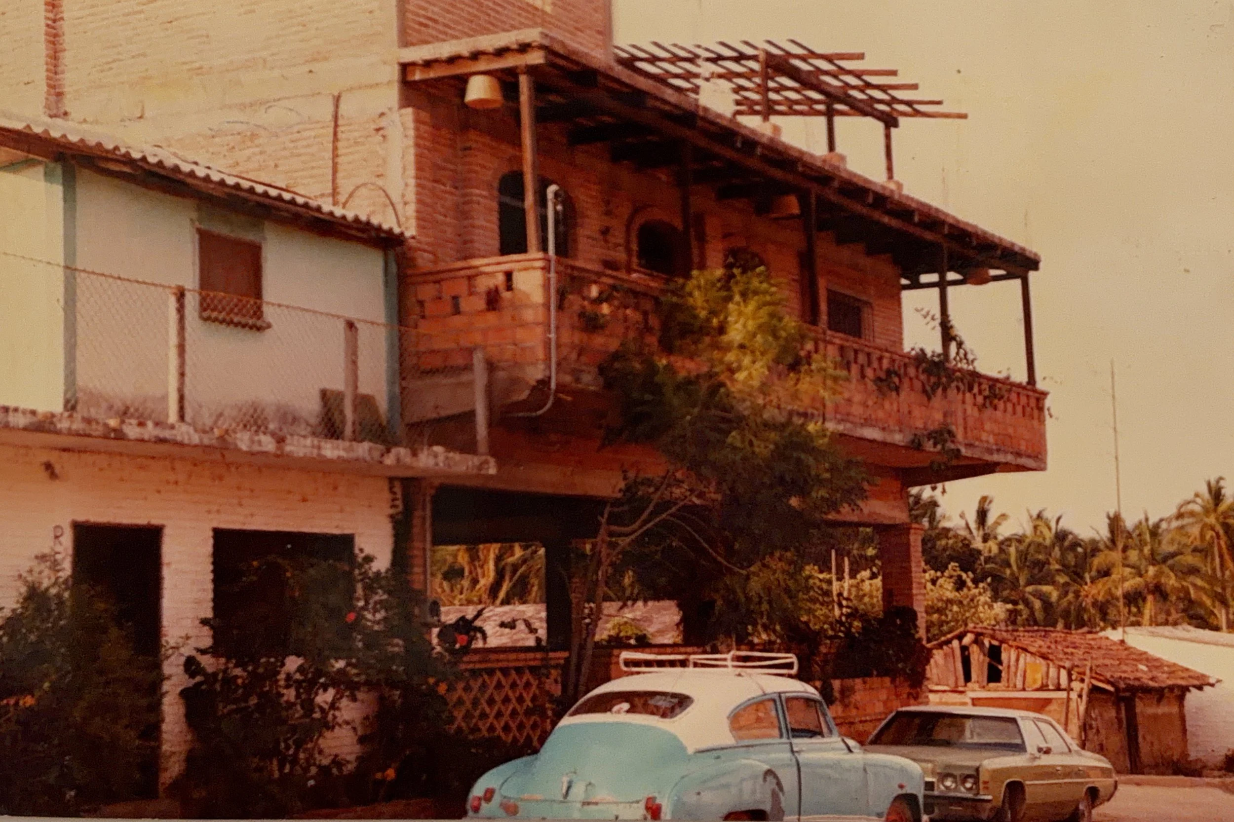 A multi-story building under construction with brick walls, a balcony, and an unfinished roof structure. Two vintage cars are parked in front, and there are trees and a fence in the background.
