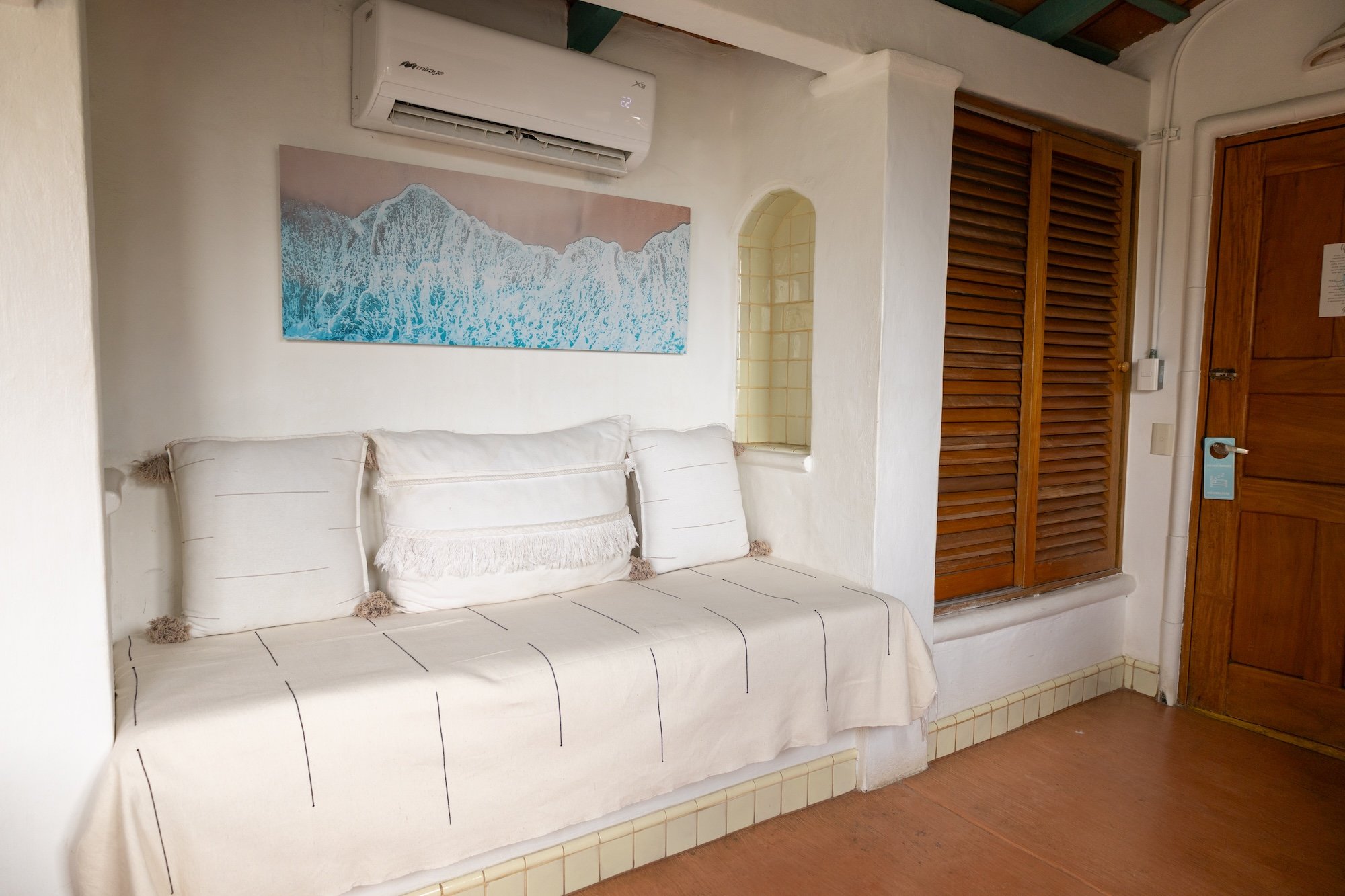 A cozy room corner with a white cushioned seating area covered by a beige fabric with black lines, topped with white pillows, a wall-mounted air conditioner above, a landscape painting of ocean waves, a small arched shelf, a louvered wooden door, and