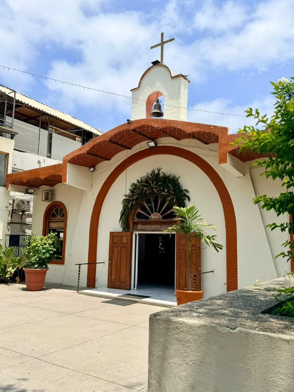 A small church with a white facade, red brick arches, and a bell tower topped with a cross. There are potted plants at the entrance and a blue sky with some clouds above.