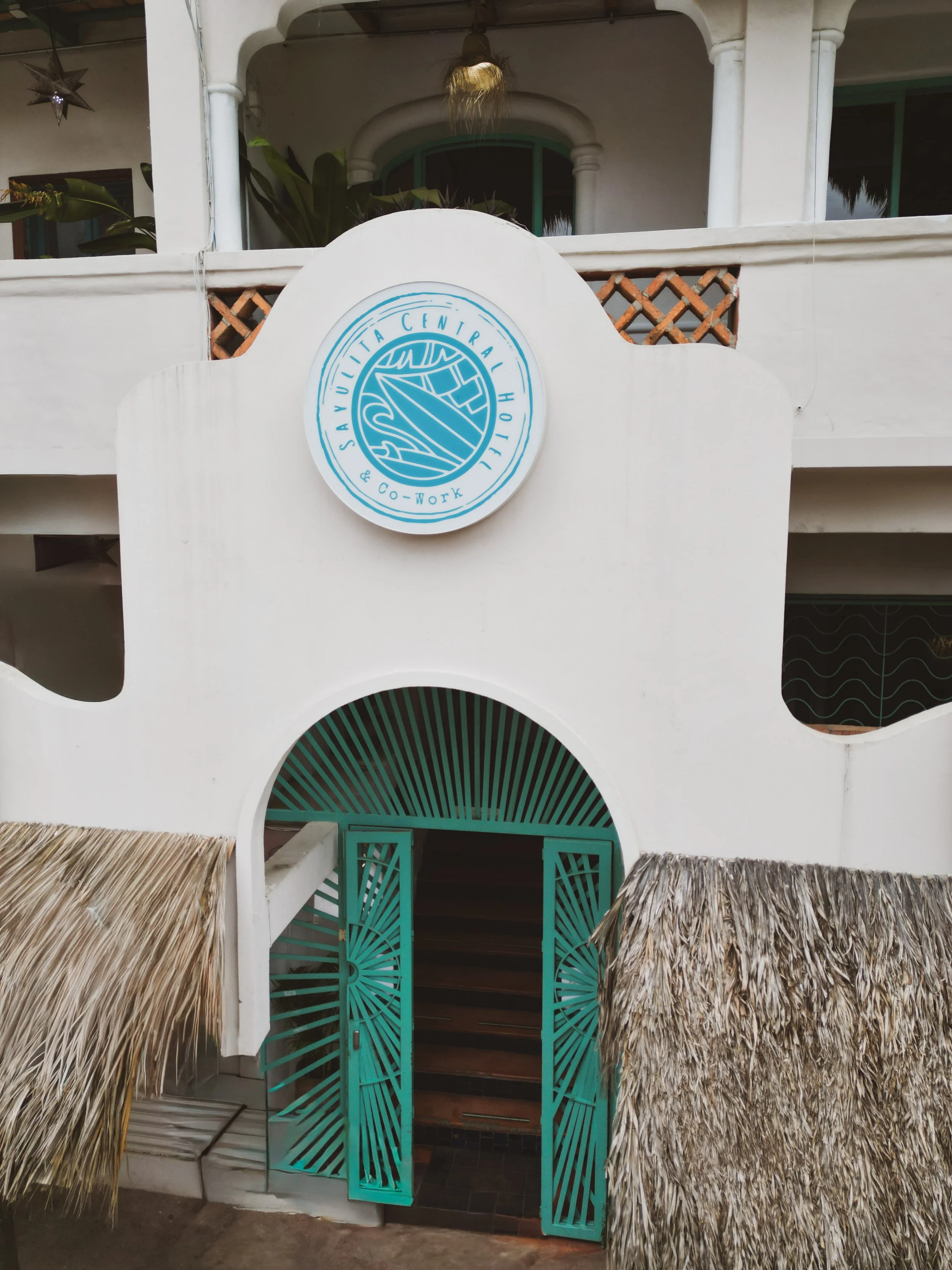 A building with a thatched roof entrance, turquoise wooden door, and a white wall with a circular logo that reads 'Savuita Central & Co-Work' with a stylized leaf design.
