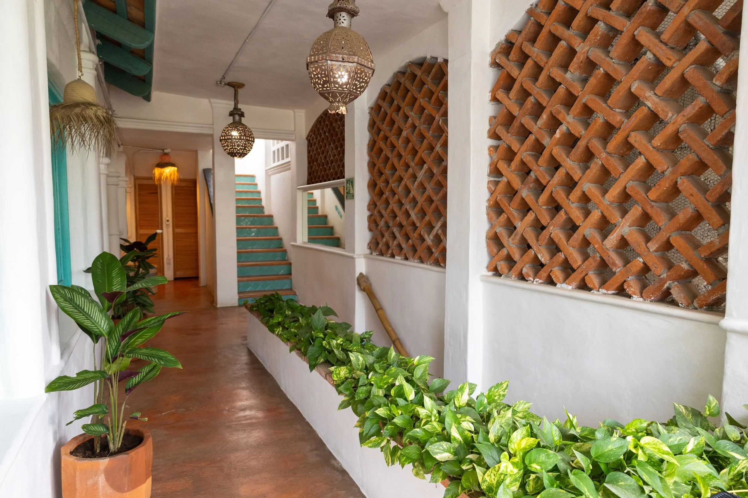 Interior hallway with green plants in pots, white walls, and a wooden floor. Decorative hanging lamps and a staircase with teal steps are visible in the background. A wall features a large brick lattice design.