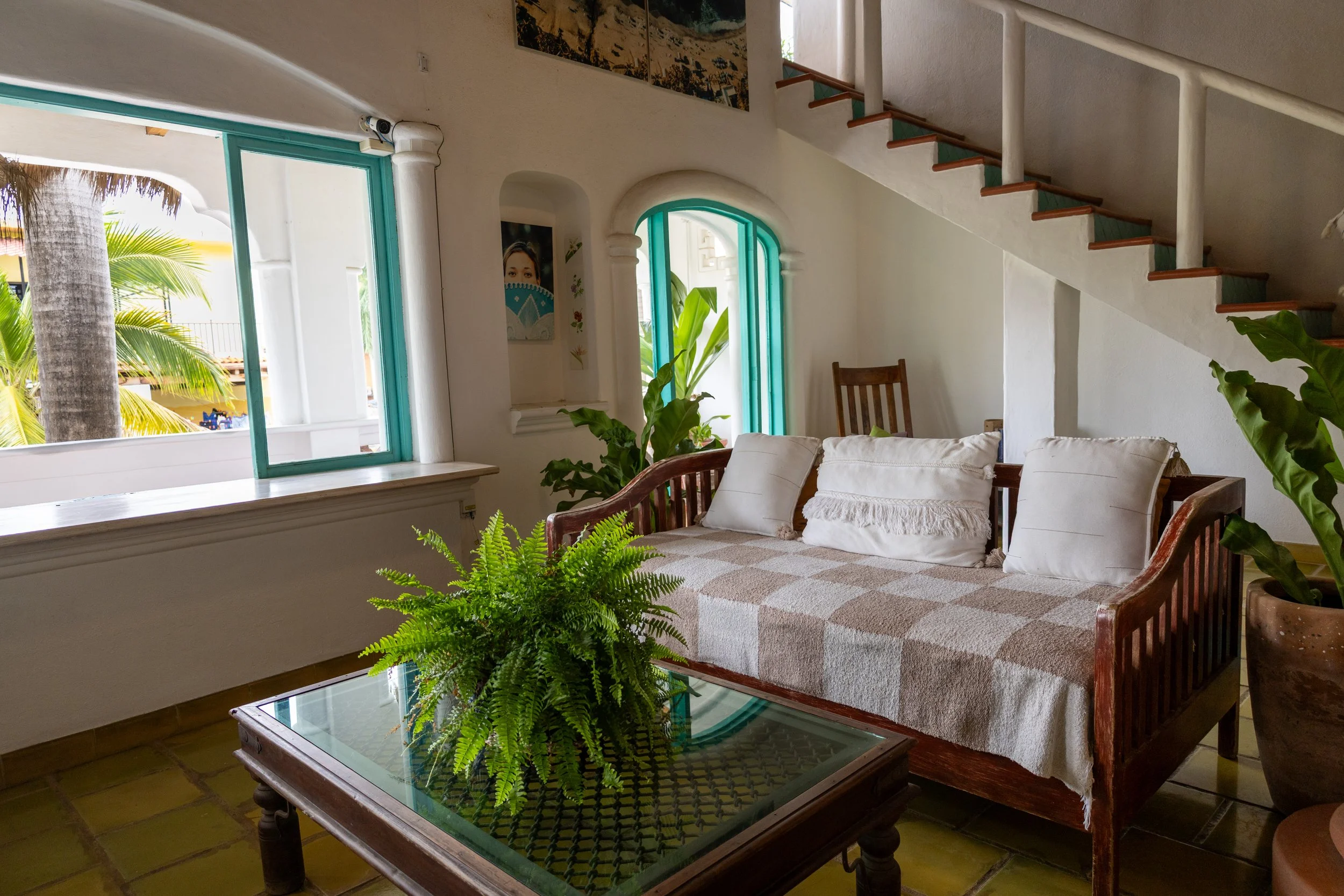 Living room with large windows showing palm trees outside, white walls, a staircase, green plants, a wooden couch with white cushions, and a glass-top coffee table with a green fern.