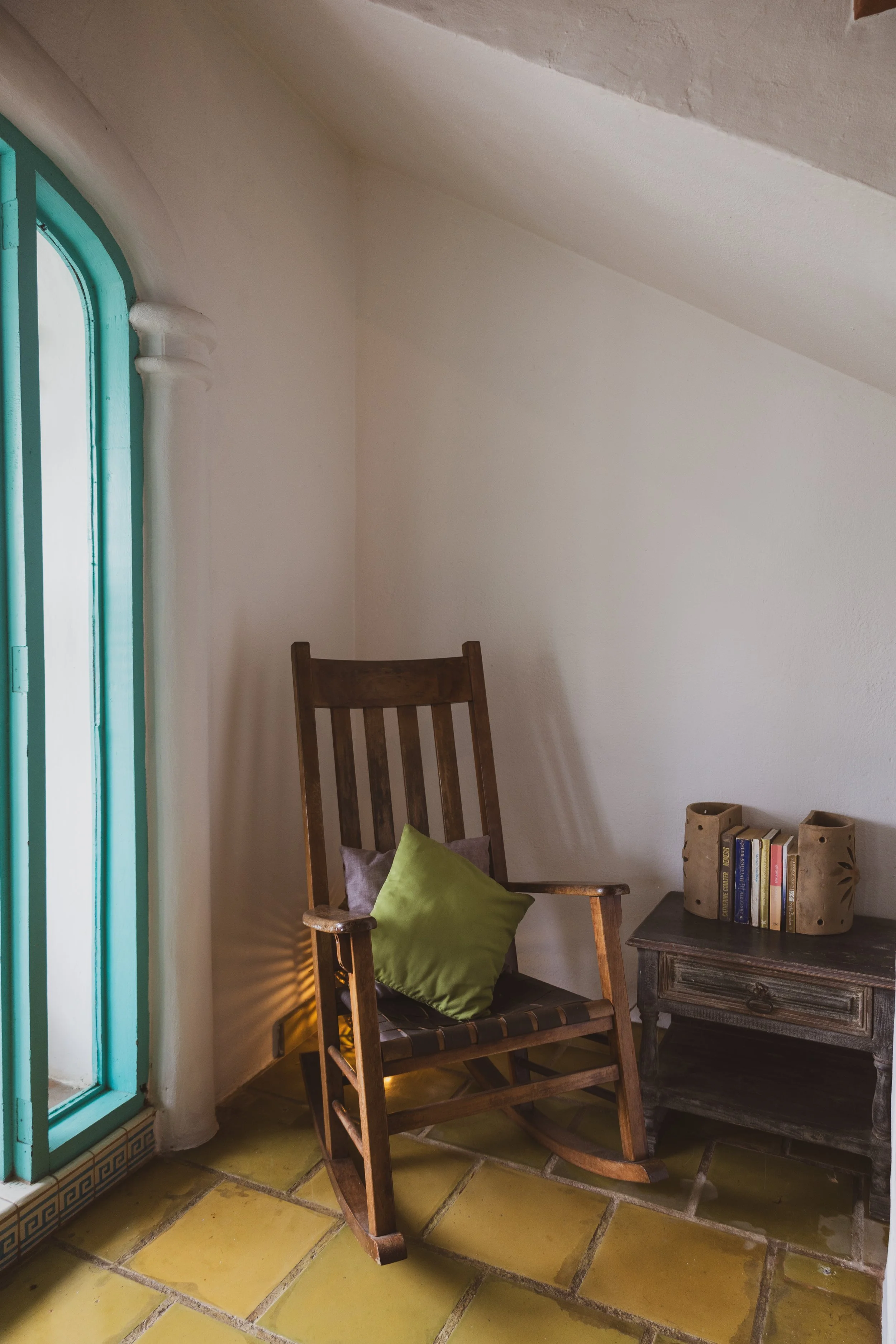 A corner of a room with a wooden chair with a green pillow, a small wooden table with books and decorative holders, and a closed door with light coming through.