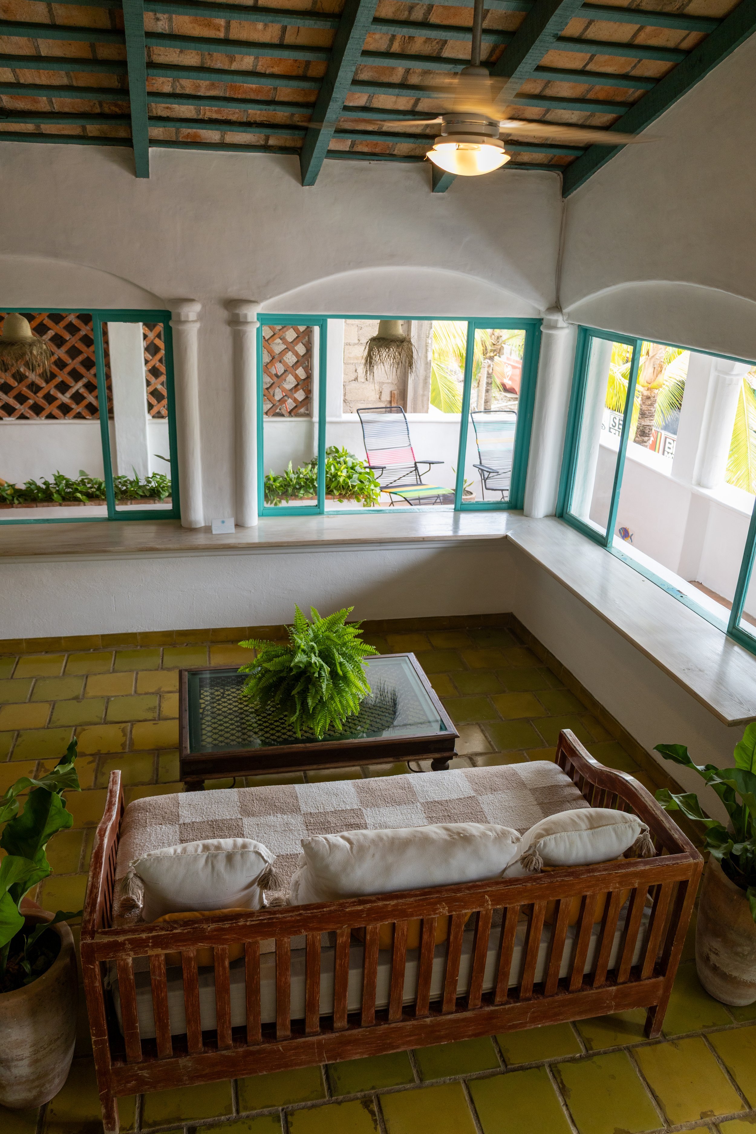 A cozy room with a tiled yellow-green floor, a wooden sofa with white cushions, potted plants, and a large window with blue-green frames showing outdoor chairs and greenery, with a ceiling fan overhead.