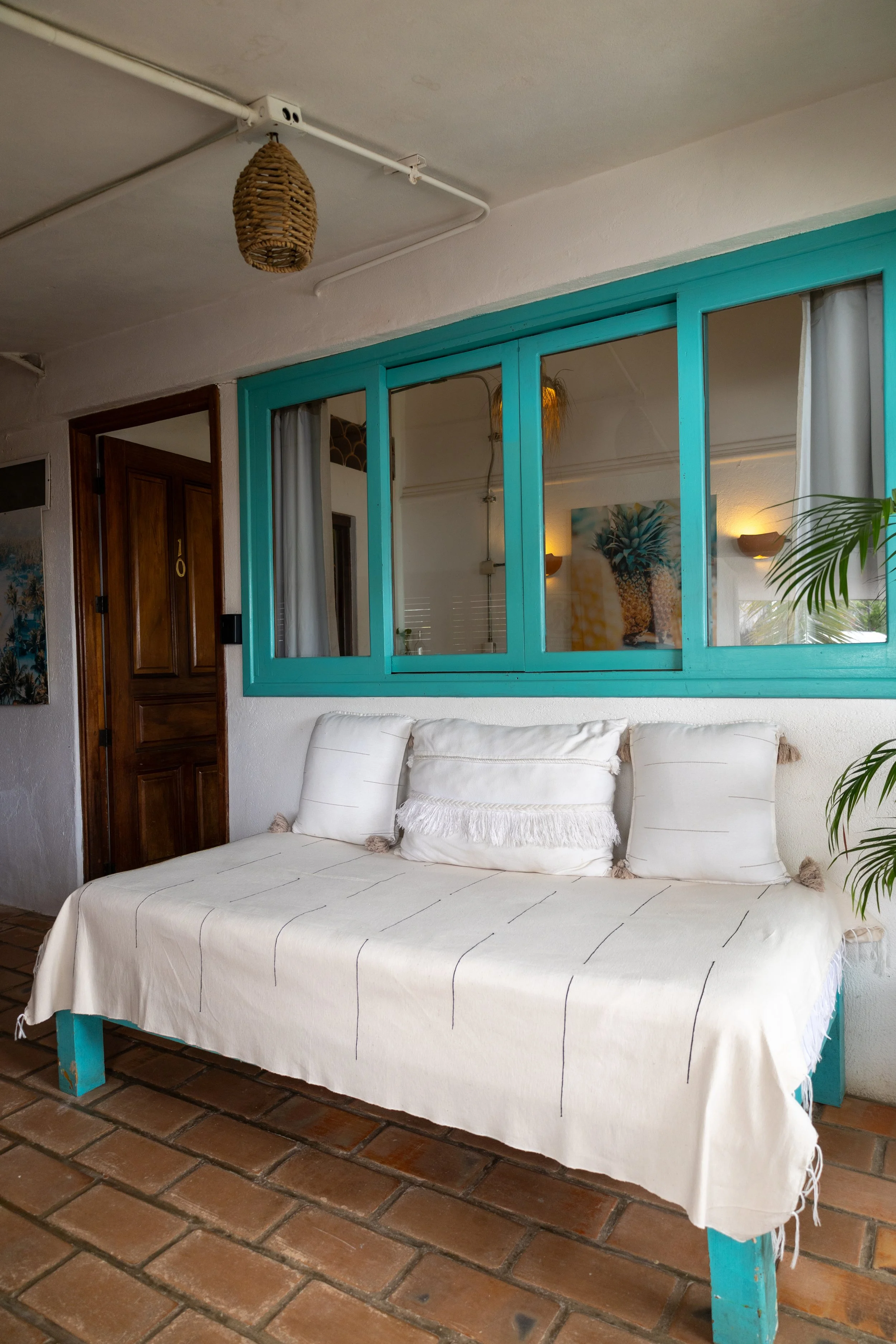 A white sitting area with a white sofa and decorative pillows, situated beneath a turquoise partition with glass windows, against a brick floor.