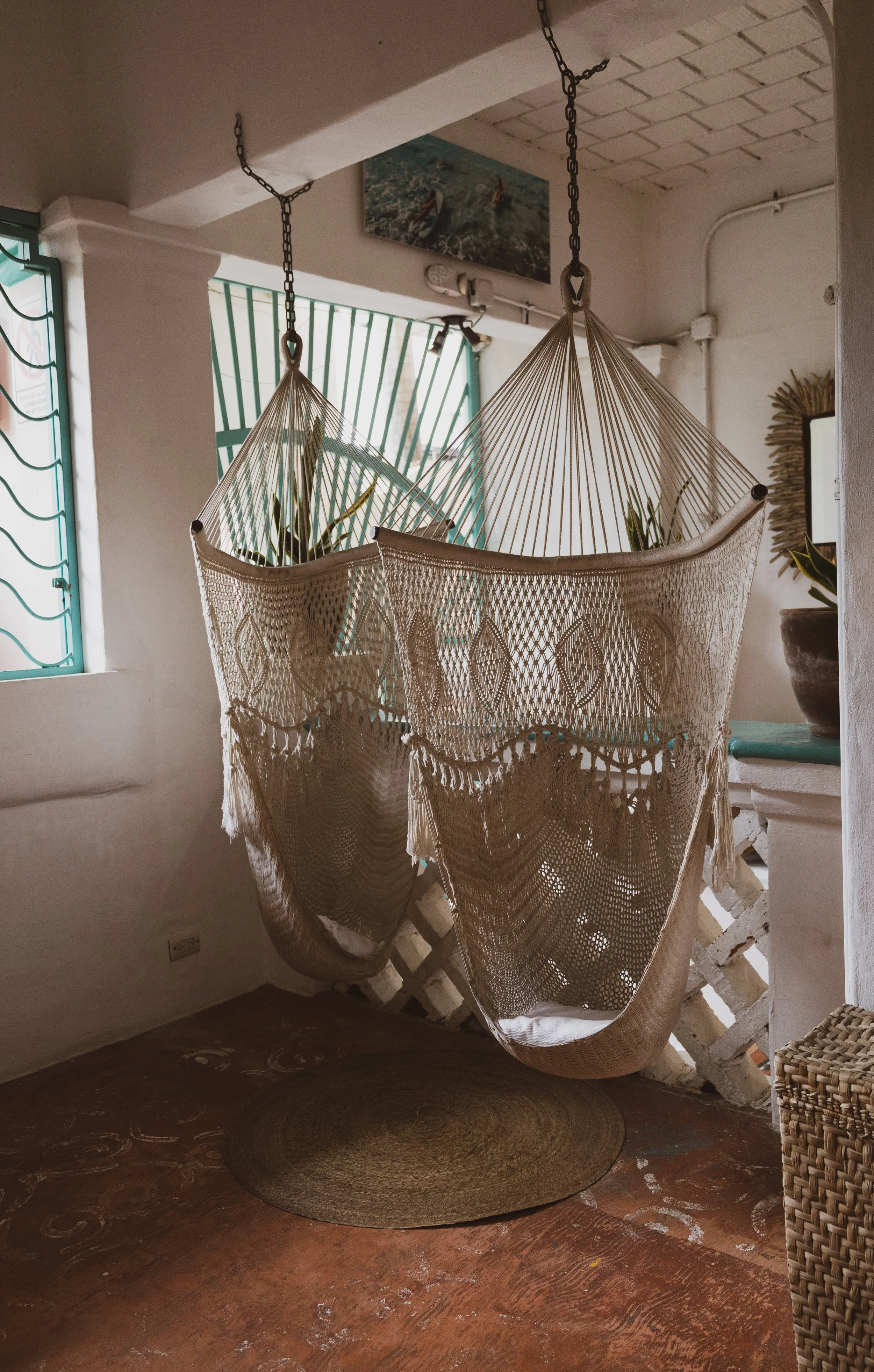 A cozy indoor space with a hanging hammock chair made of woven fabric and strings, suspended from the ceiling with chains. There is a small round rug on the wooden floor underneath the hammock. A window with green metal bars, a framed picture of surf