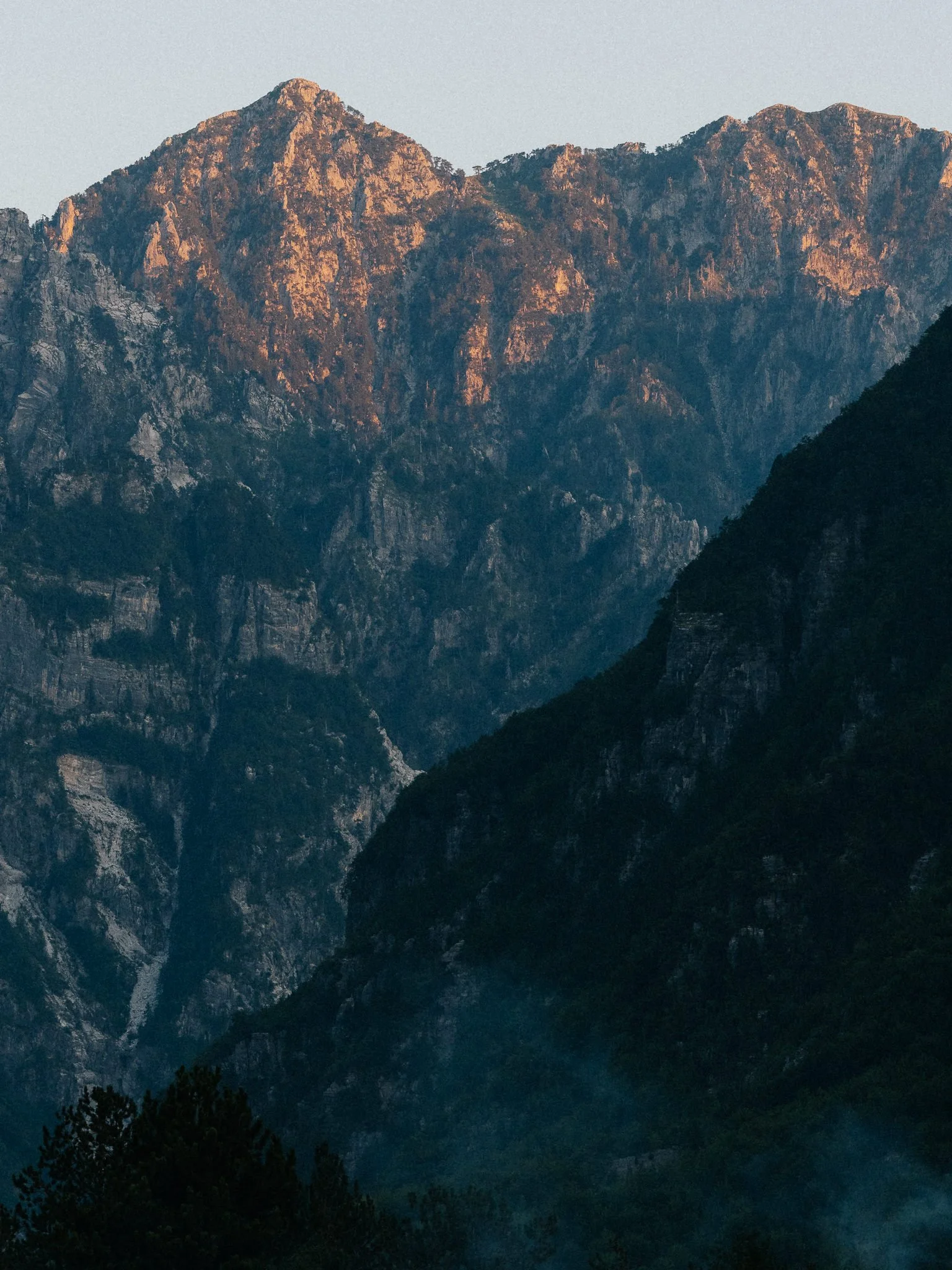 Mountain range at dusk with rugged, rocky peaks illuminated by setting sun, framed by dark trees in the foreground.