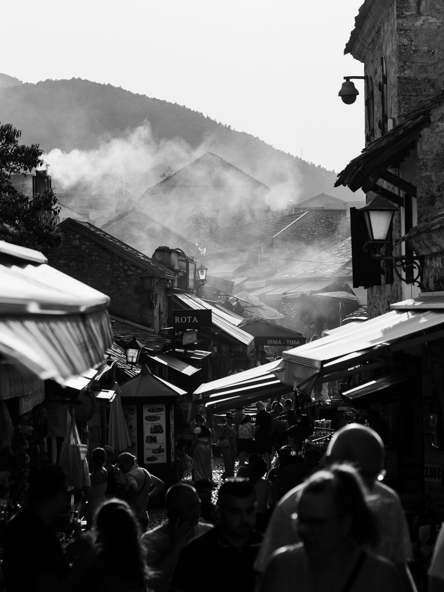 Crowded outdoor market scene with shop stalls, umbrellas, and blurred people walking, mountains and smoke in the background, black and white photo.