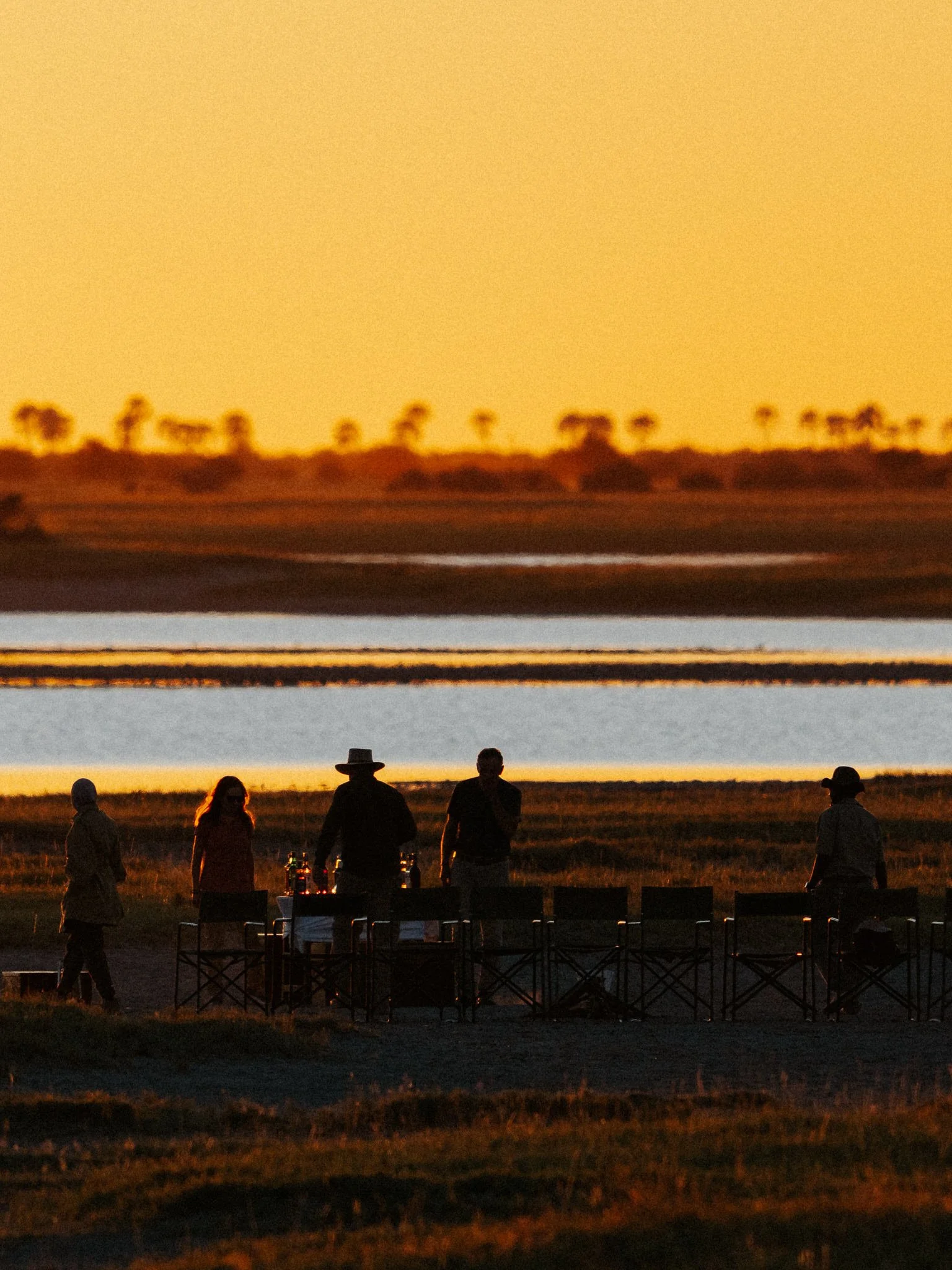 Group of people standing around a table outdoors during sunset, with a distant landscape of trees and a body of water.