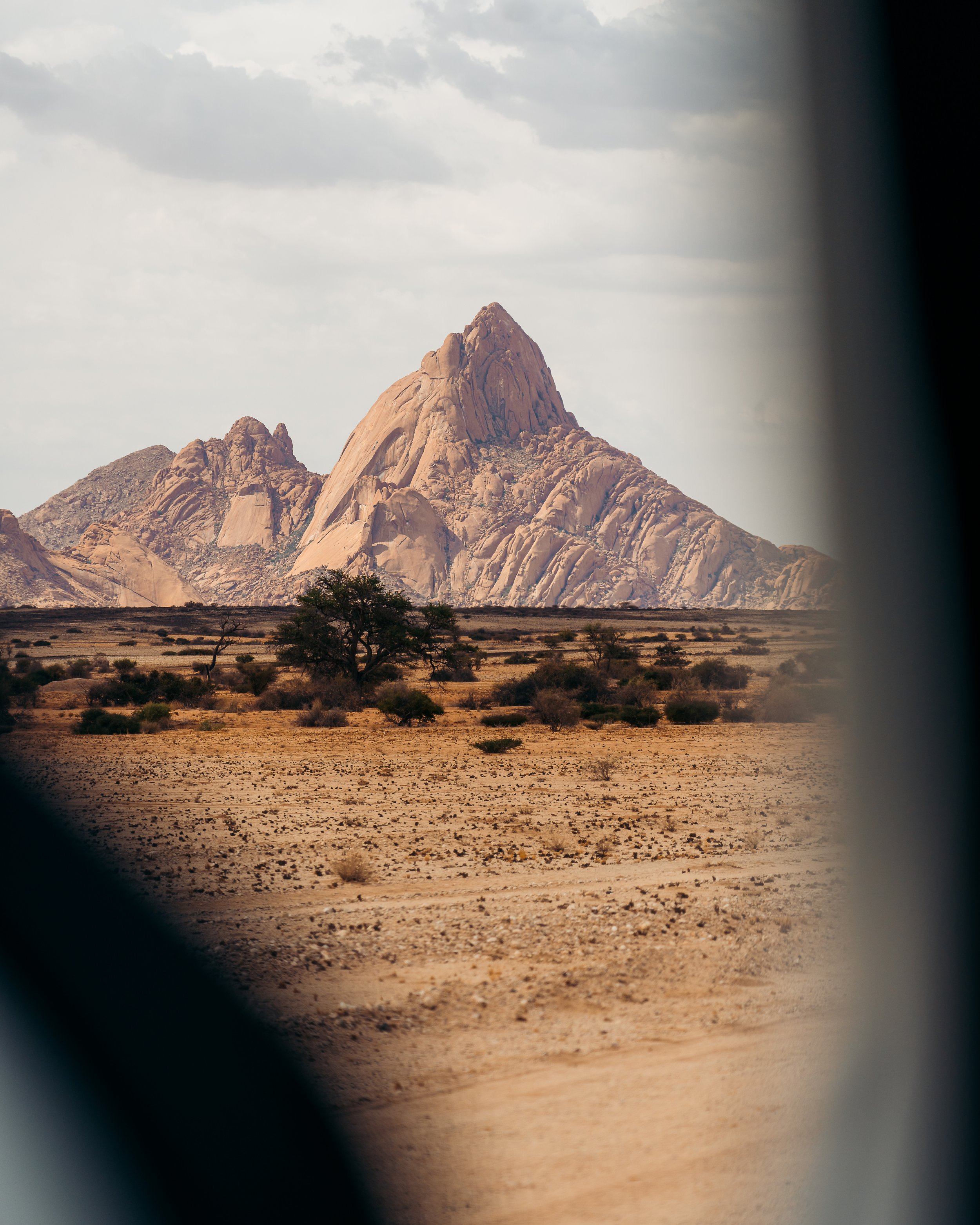 Desert landscape with large rocky mountain and sparse vegetation, viewed through a window or opening.