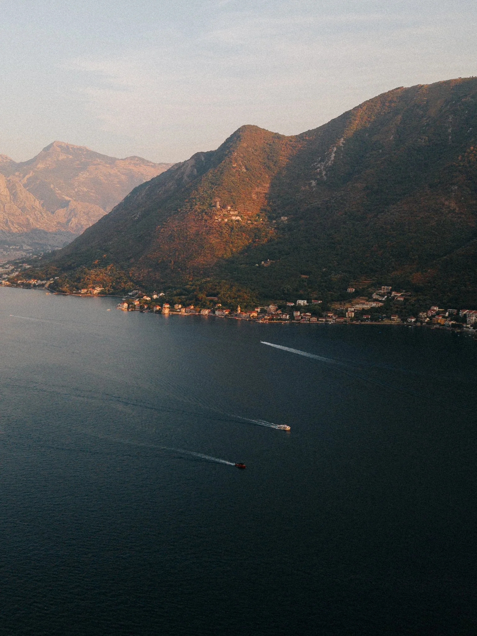 A large lake surrounded by mountains with a small town at the shoreline, and two boats creating wakes on the water.