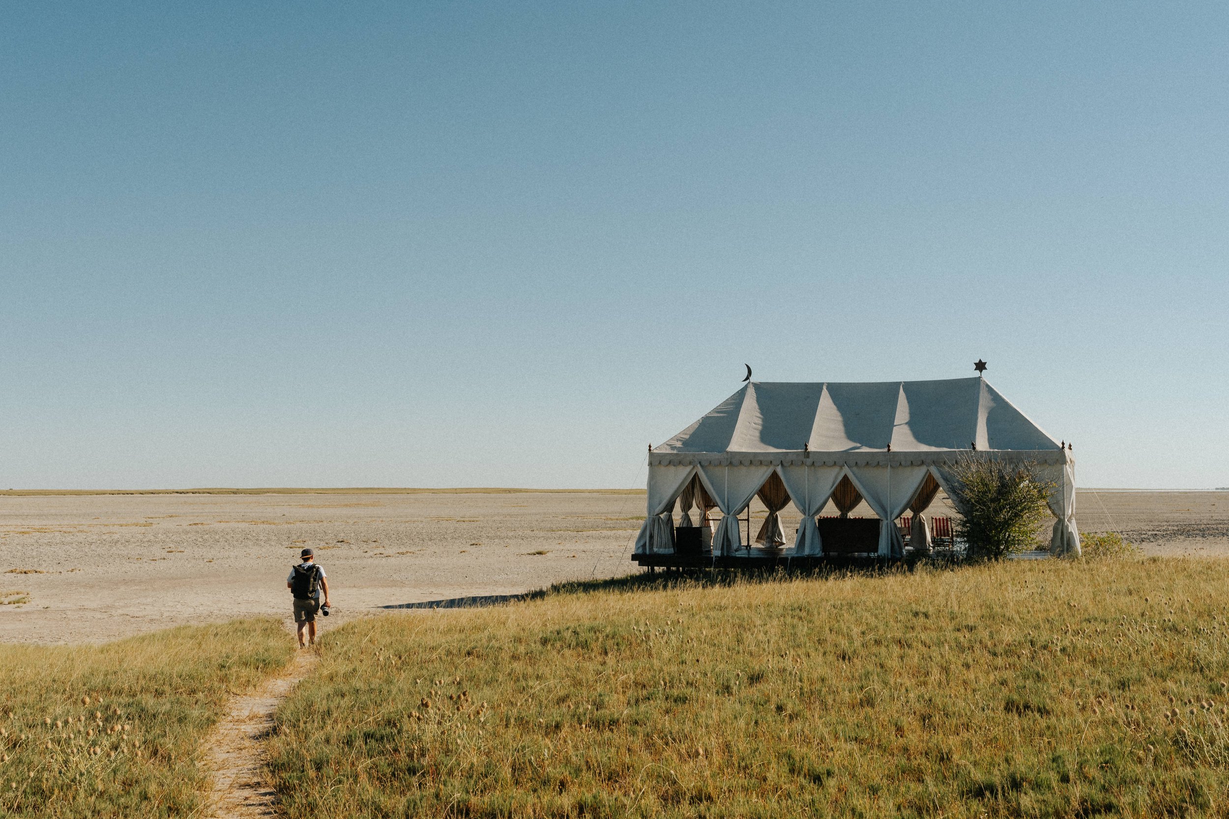 A person with a backpack walking towards a large white circus tent in an open field under a clear blue sky.